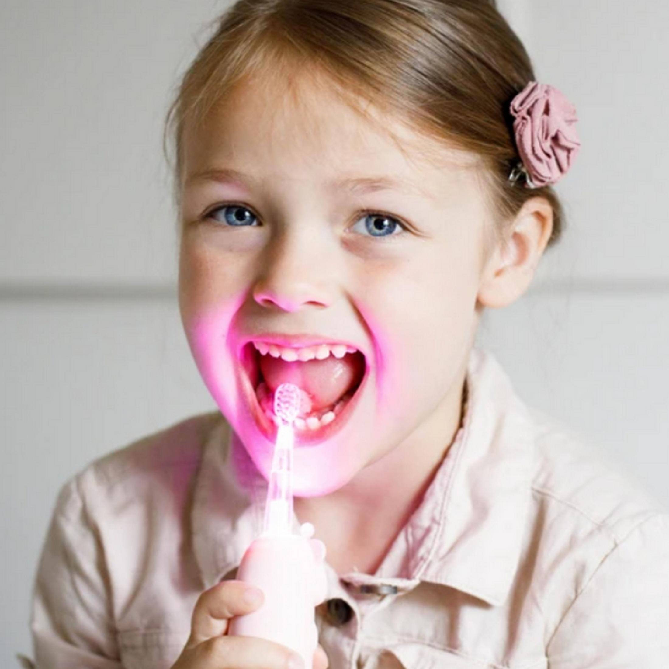 Una bambina con un accessorio rosa tra i capelli si lava i denti con uno spazzolino rosa luminoso.