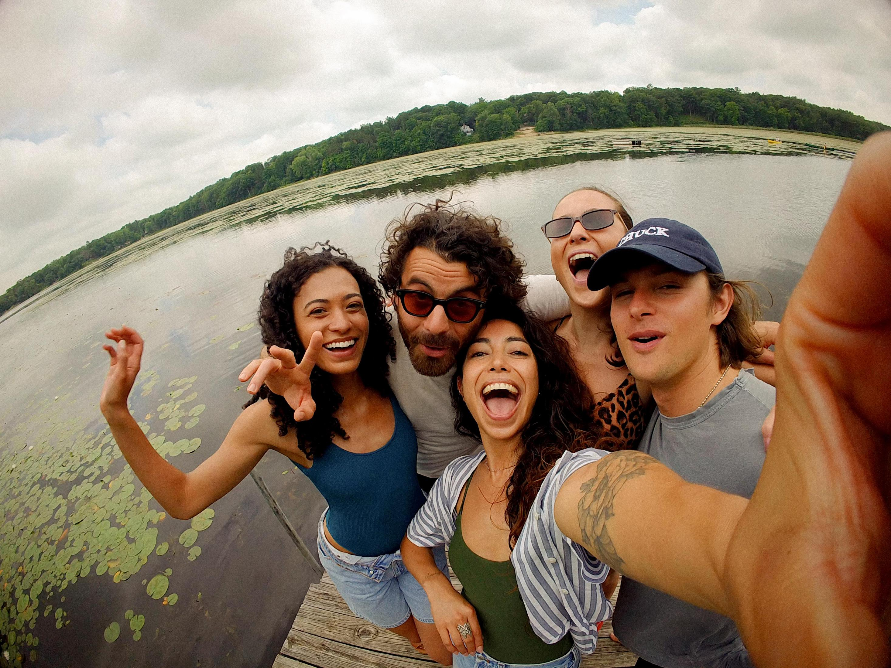 Cinque amici si scattano un selfie su un molo di legno vicino a un lago con ninfee e alberi.