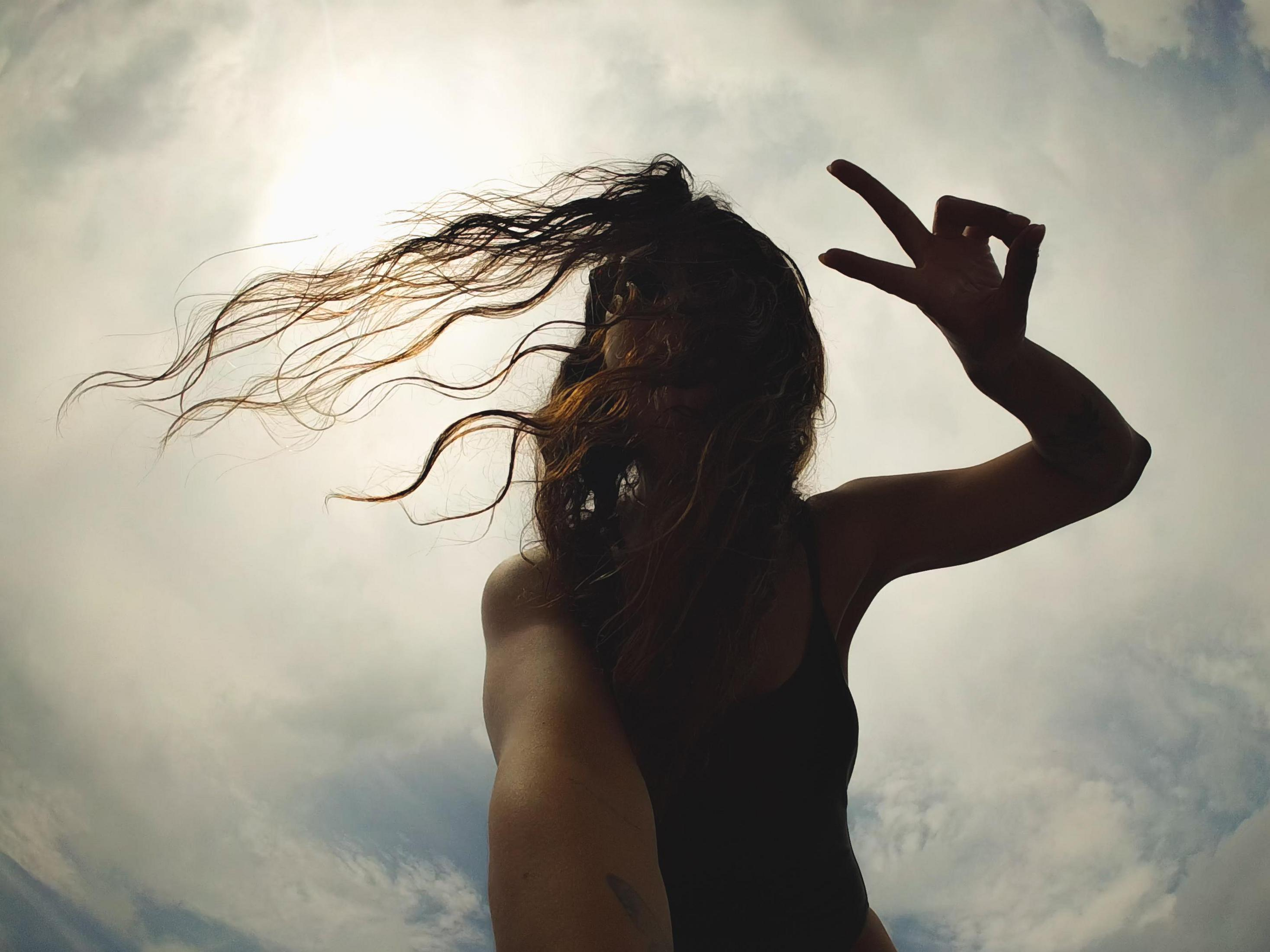 Silhouette di una donna con capelli al vento che fa il segno a V contro un cielo nuvoloso.