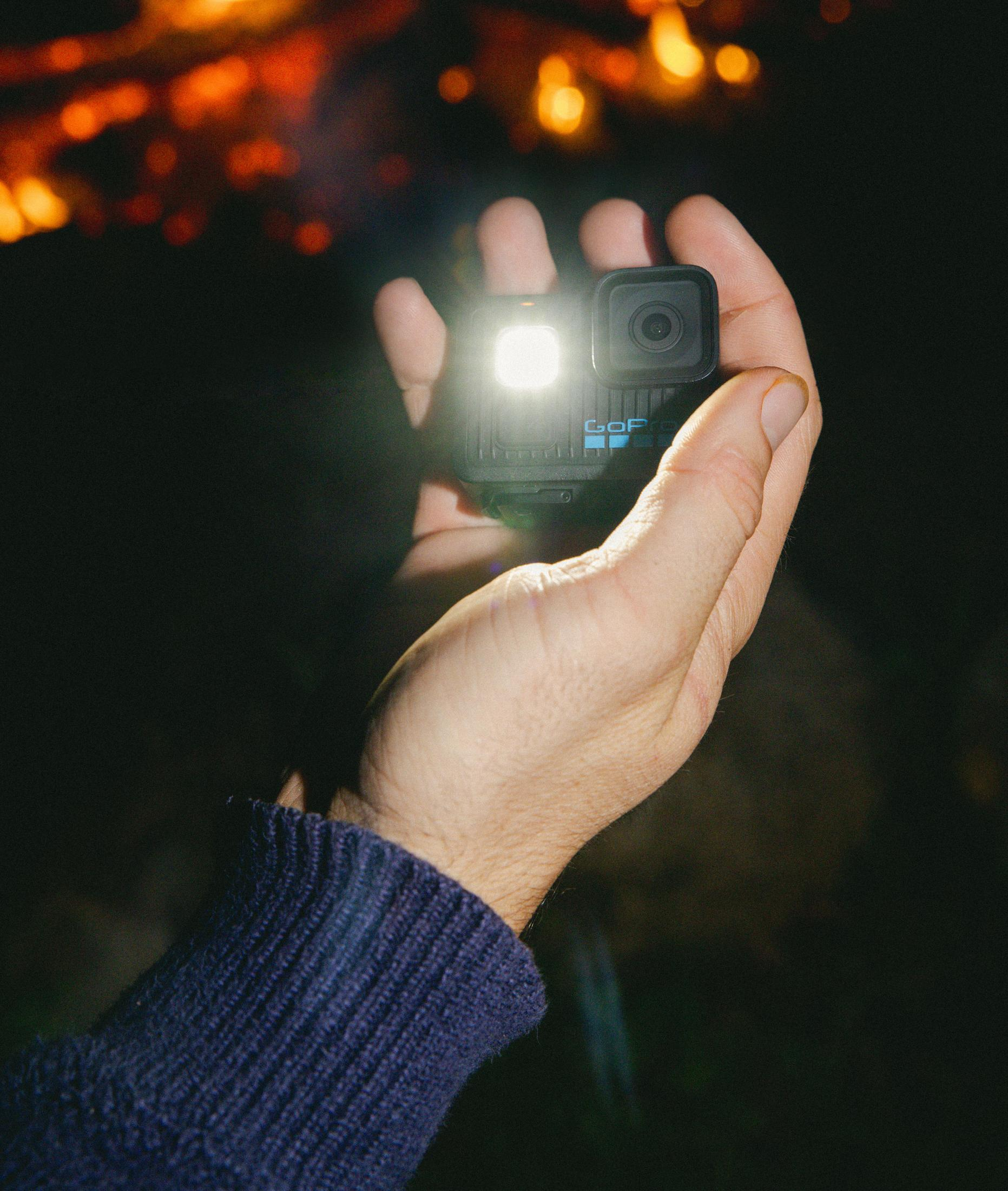 Una mano tiene una GoPro con la luce accesa, davanti a un fuoco sfocato.