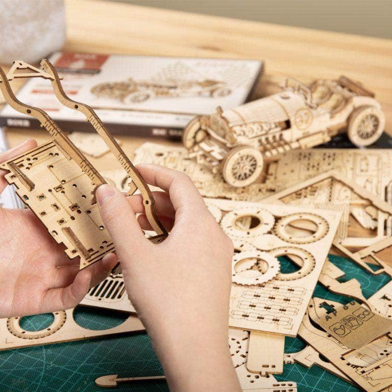 Hände bauen ein Holzauto-Modellbausatz. Viele Holzteile und ein Modellauto sind sichtbar.