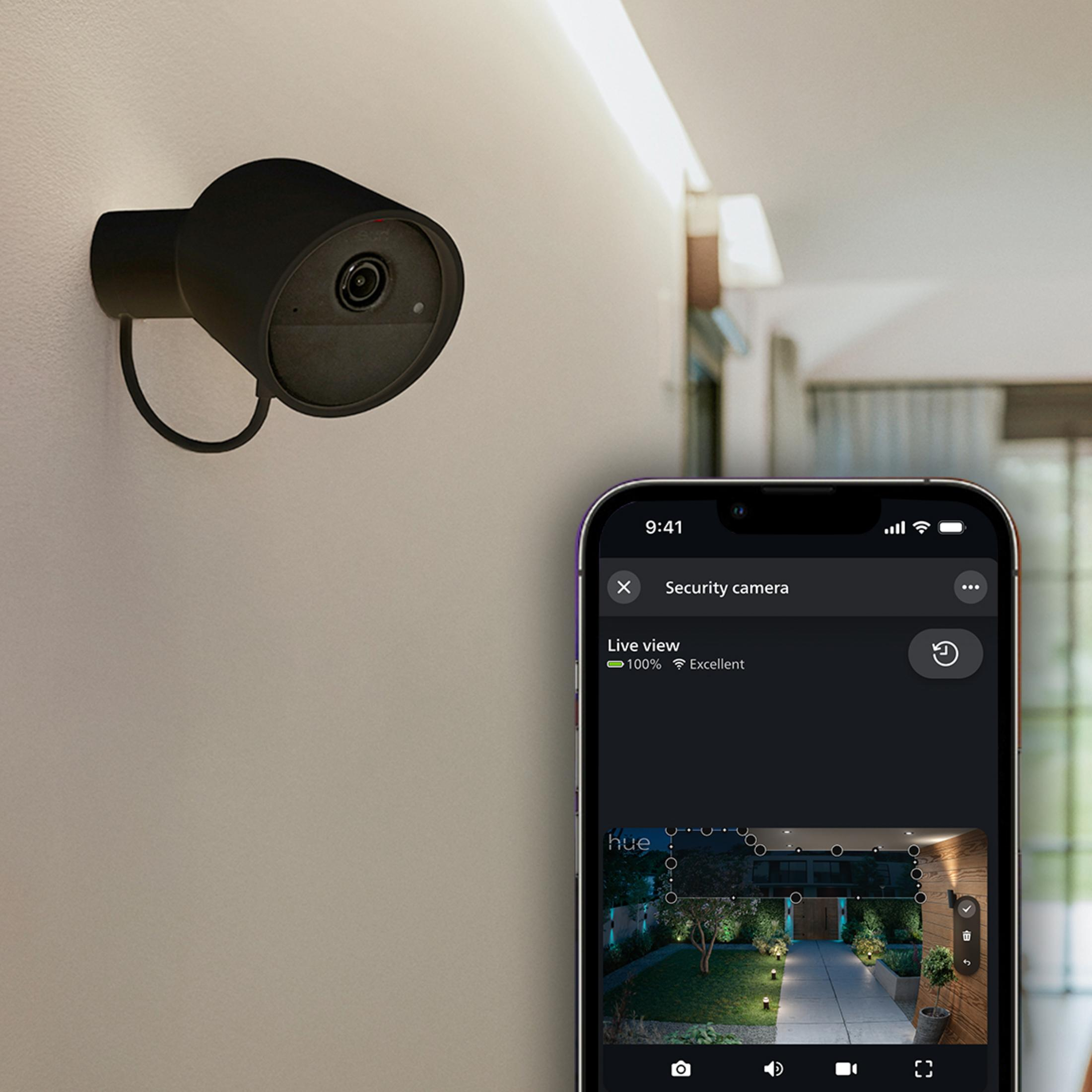 Una cámara de seguridad negra en una pared blanca, junto a un smartphone con vista en vivo del jardín.