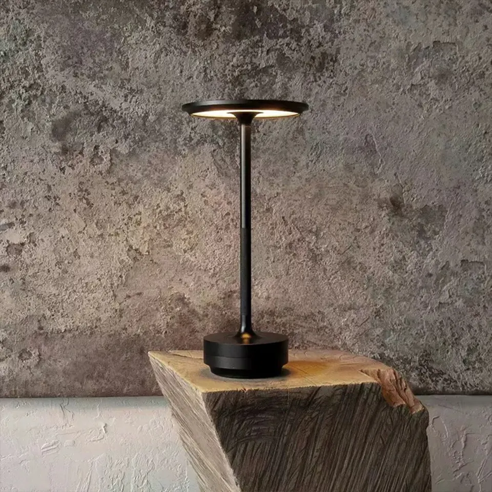 Eine schwarze Tischlampe mit rundem Lampenschirm auf einem Holzblock vor einer strukturierten Wand.