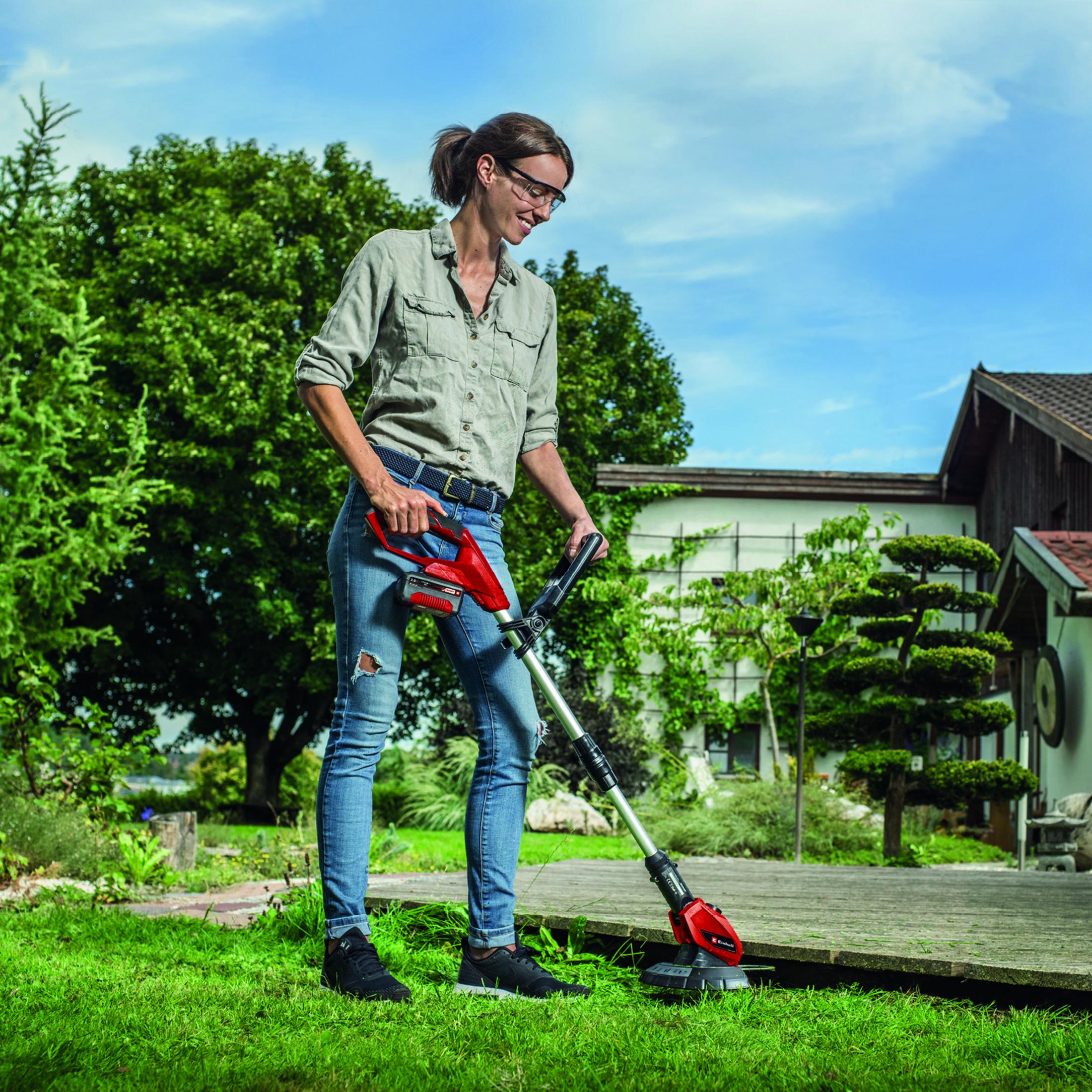 Frau trimmt Rasen mit einem rot-schwarzen Trimmer auf einer Holzterrasse im Garten.