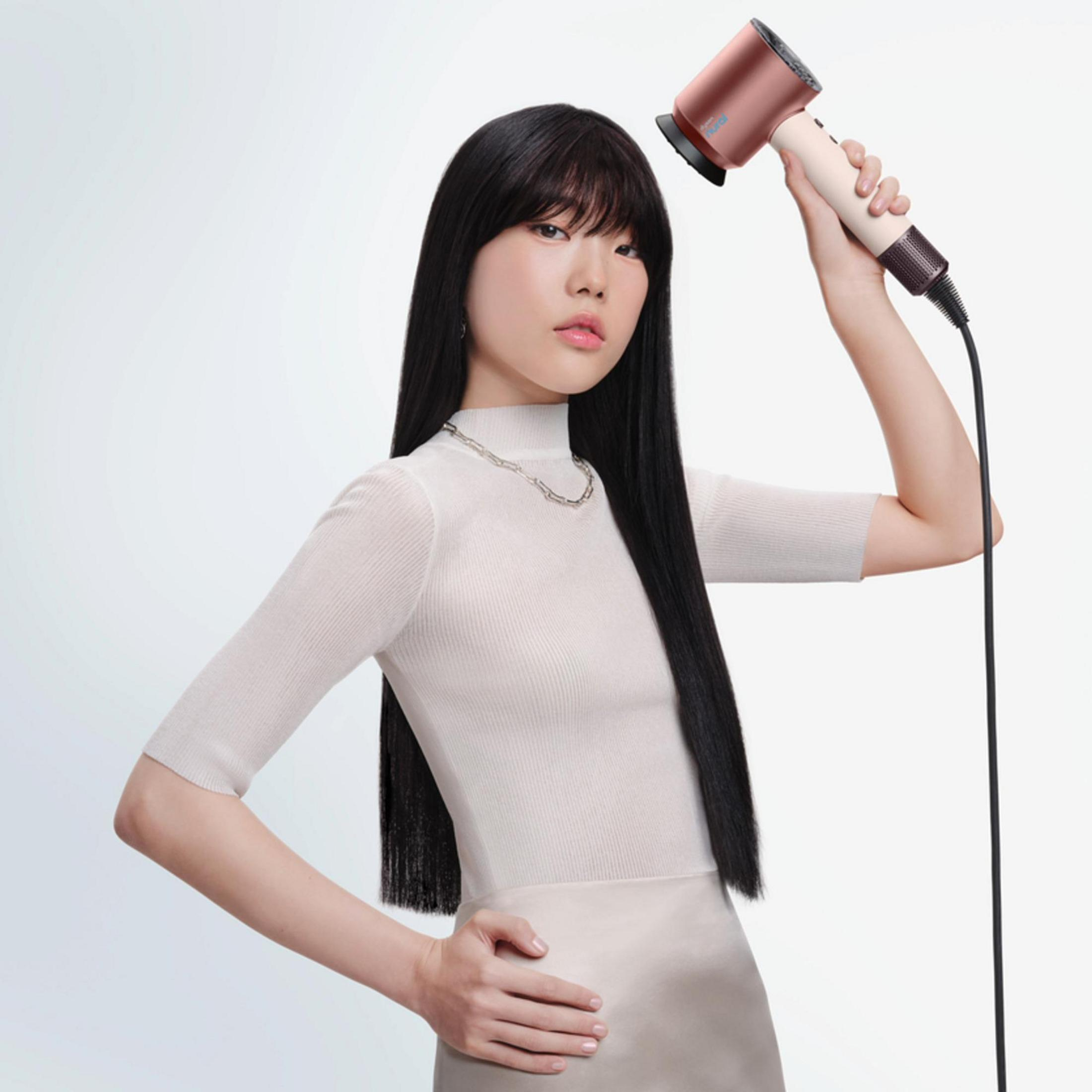 Eine Frau mit langen schwarzen Haaren hält einen rosafarbenen Haartrockner über ihren Kopf.