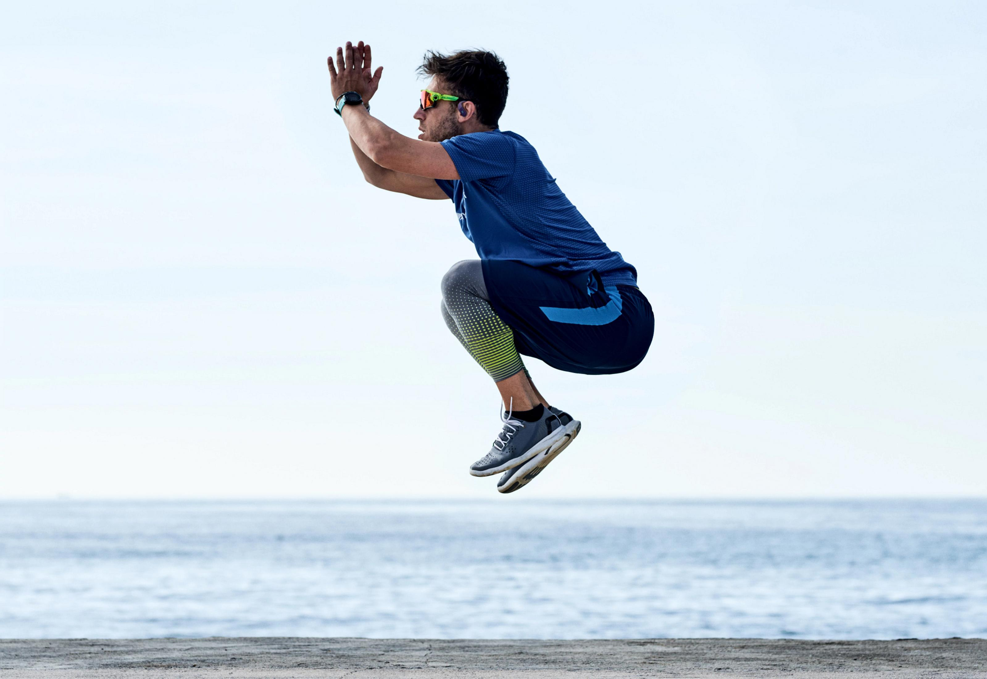 Un uomo in abbigliamento sportivo salta sul mare, mani alzate.
