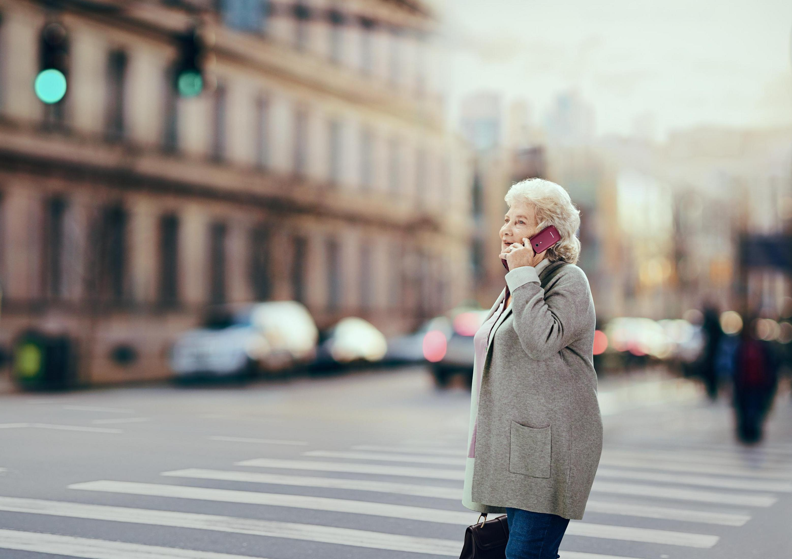 Un'anziana parla al telefono rosa su un attraversamento pedonale. Strada cittadina con edifici e auto sfocati.