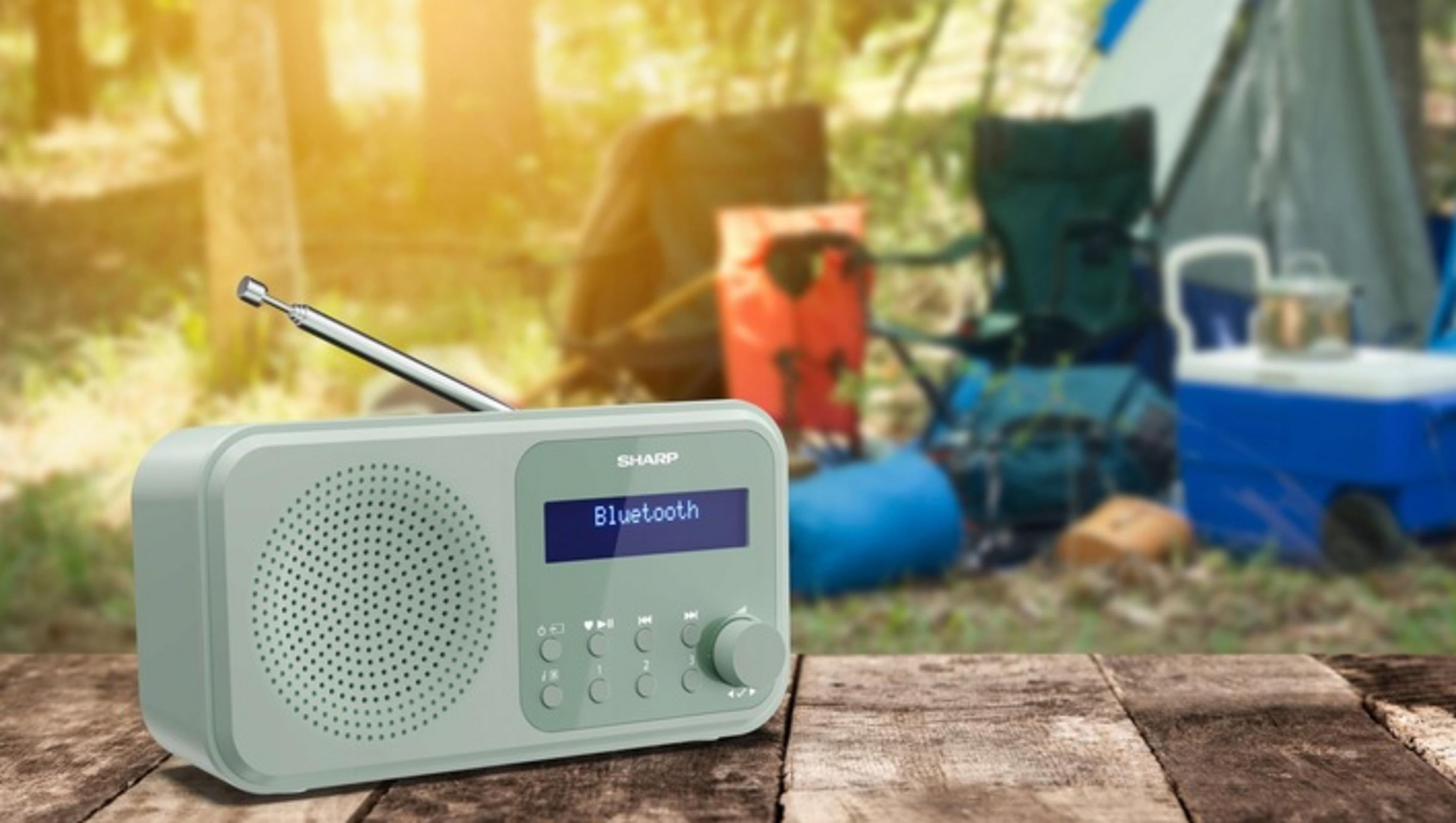 Ein mintgrünes Radio mit Bluetooth steht auf einem Holztisch, Campingausrüstung im Hintergrund.