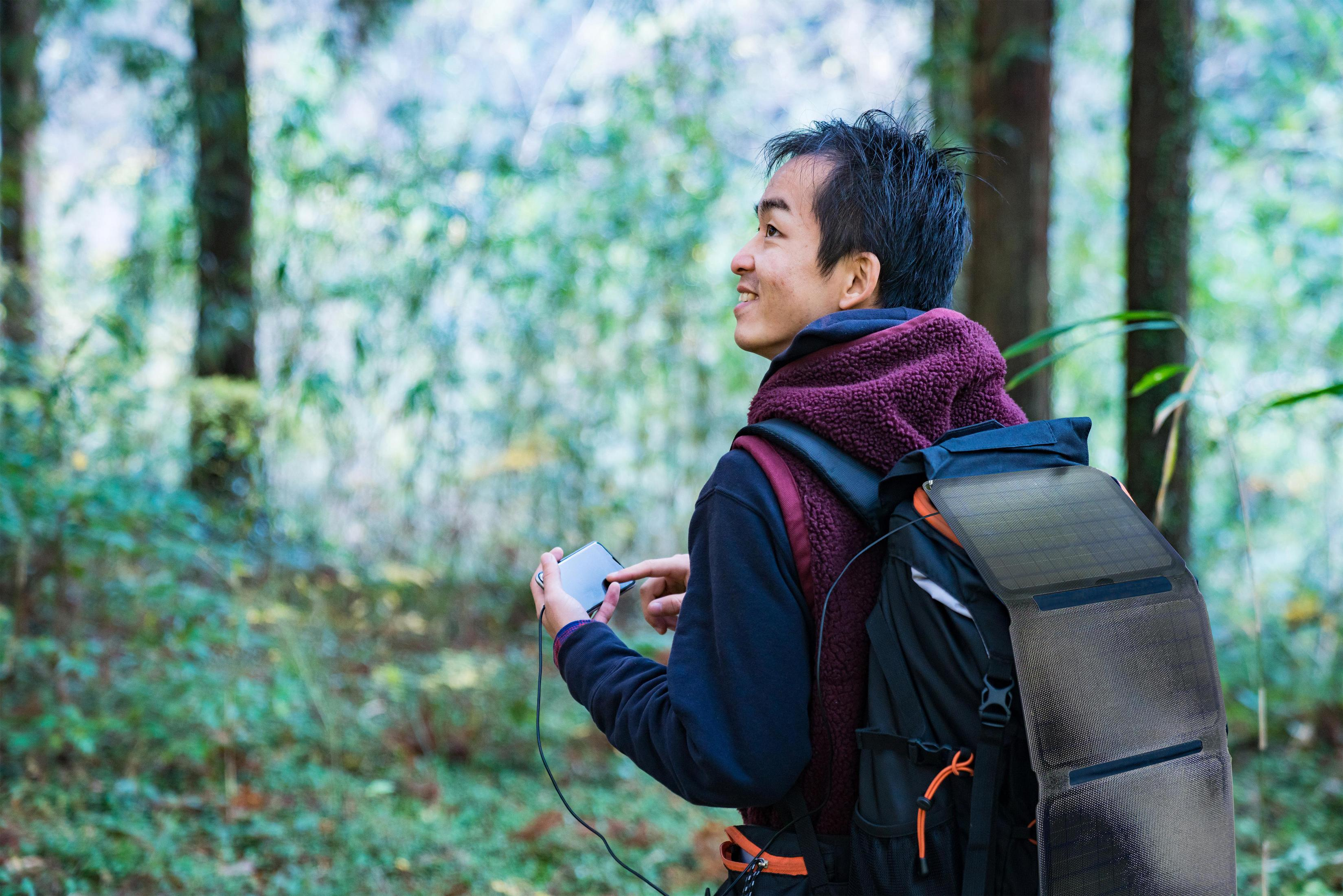 Un uomo con zaino e pannello solare in una foresta, guarda il suo telefono.