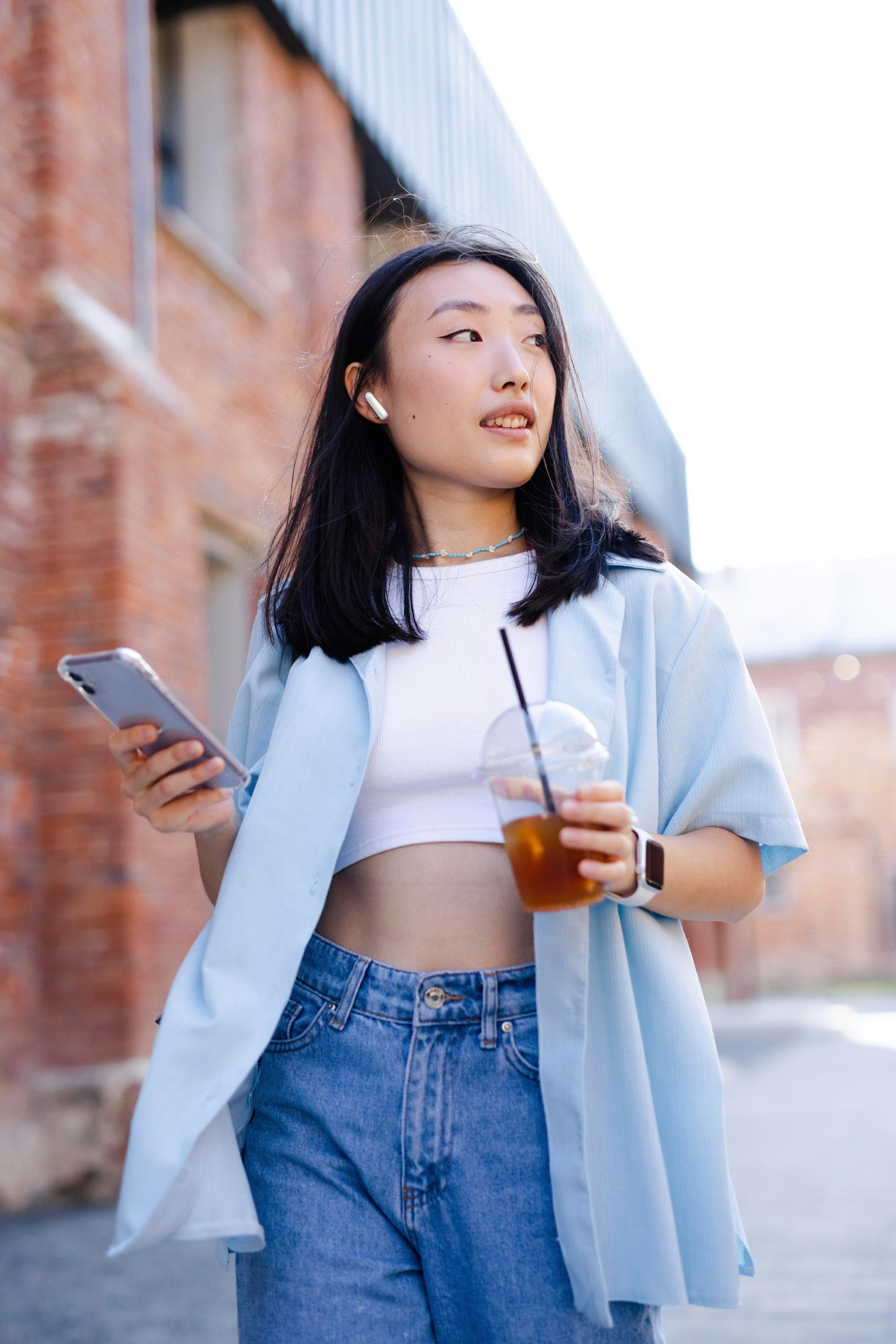 Una giovane donna con auricolari tiene un telefono e una bevanda. Indossa jeans e una camicia azzurra.