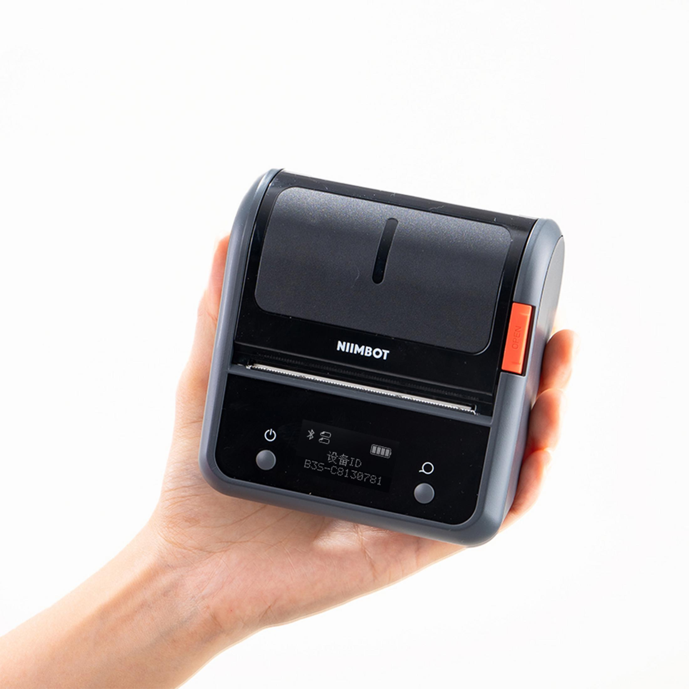 Una mano tiene una stampante portatile NIIMBOT grigio scuro con pulsante arancione 'OPEN'.