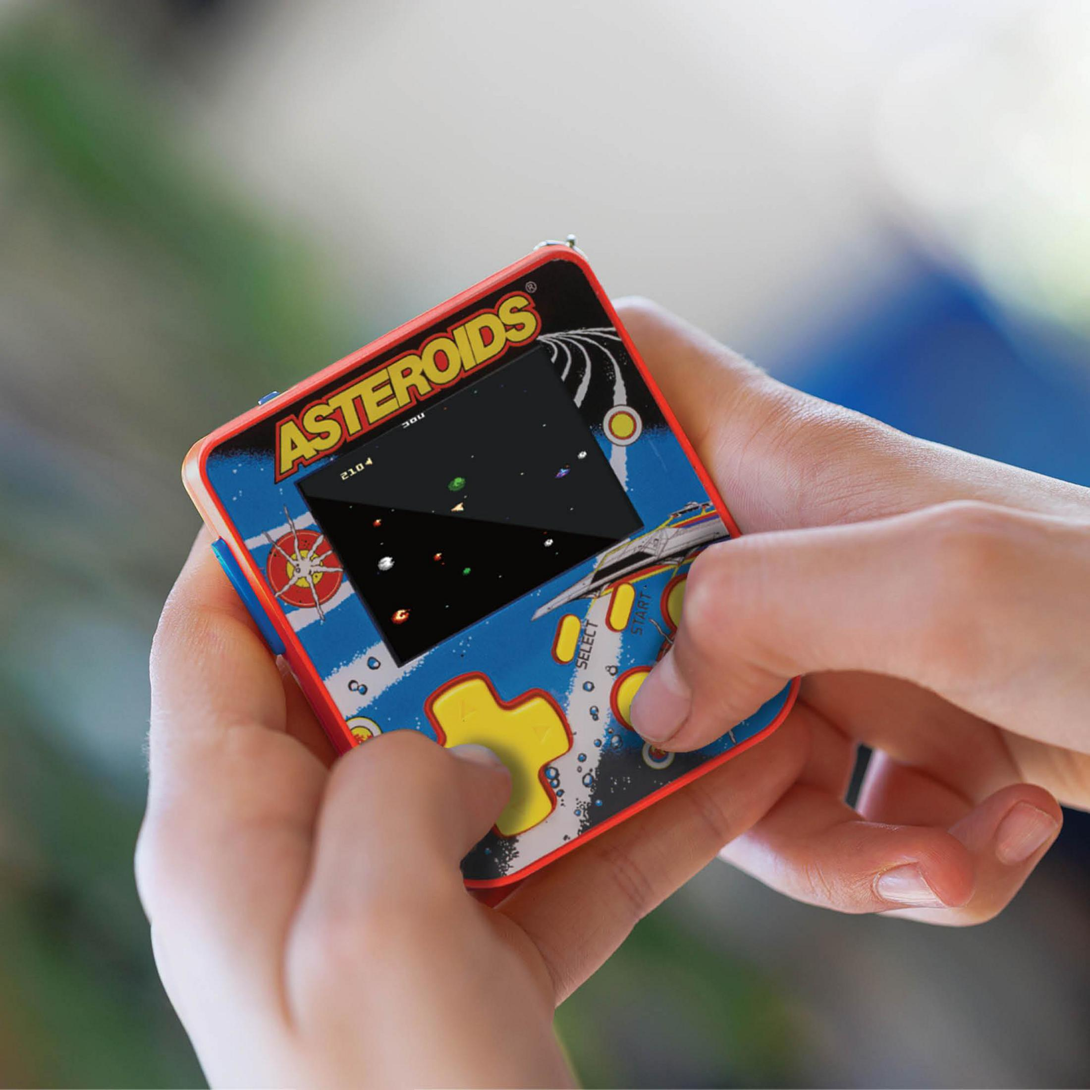 Manos sostienen un juego arcade de mano con tema de Asteroides. Un pulgar presiona un botón.