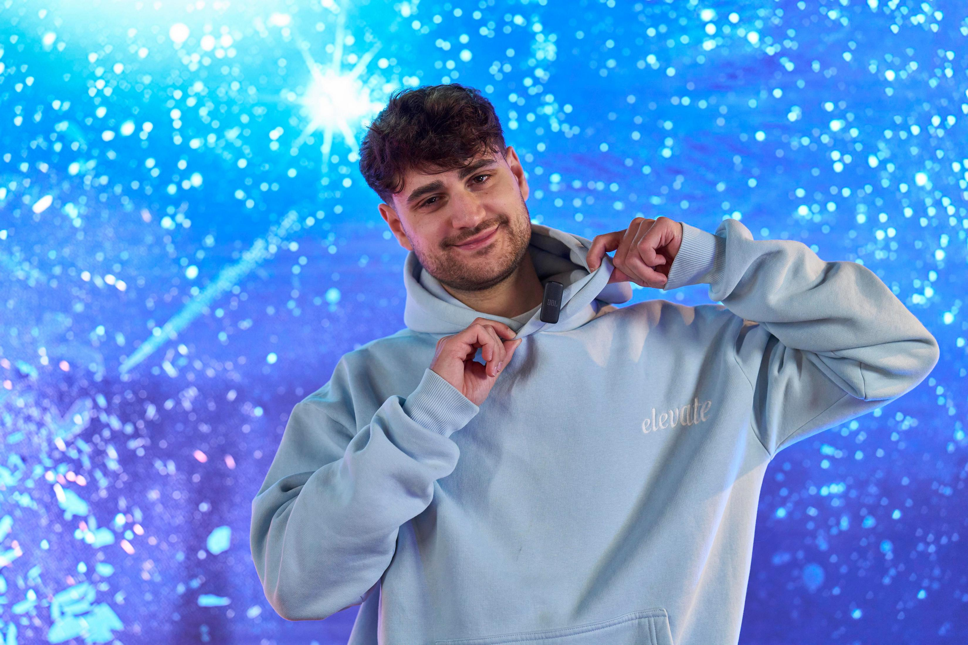 Ein Mann mit lockigem Haar trägt einen hellblauen Hoodie vor einem blauen, funkelnden Hintergrund.