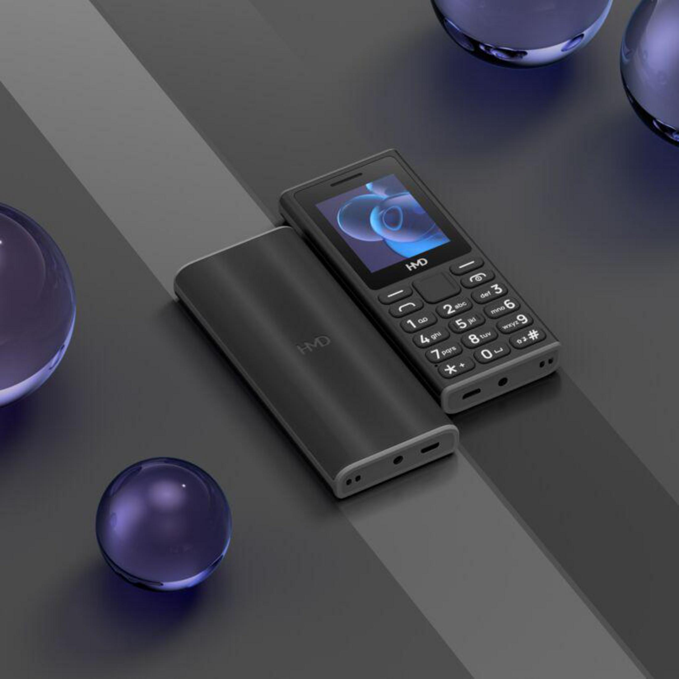 Ein schwarzes Handy mit Tasten und ein zweites Gerät liegen auf einem grauen Untergrund.