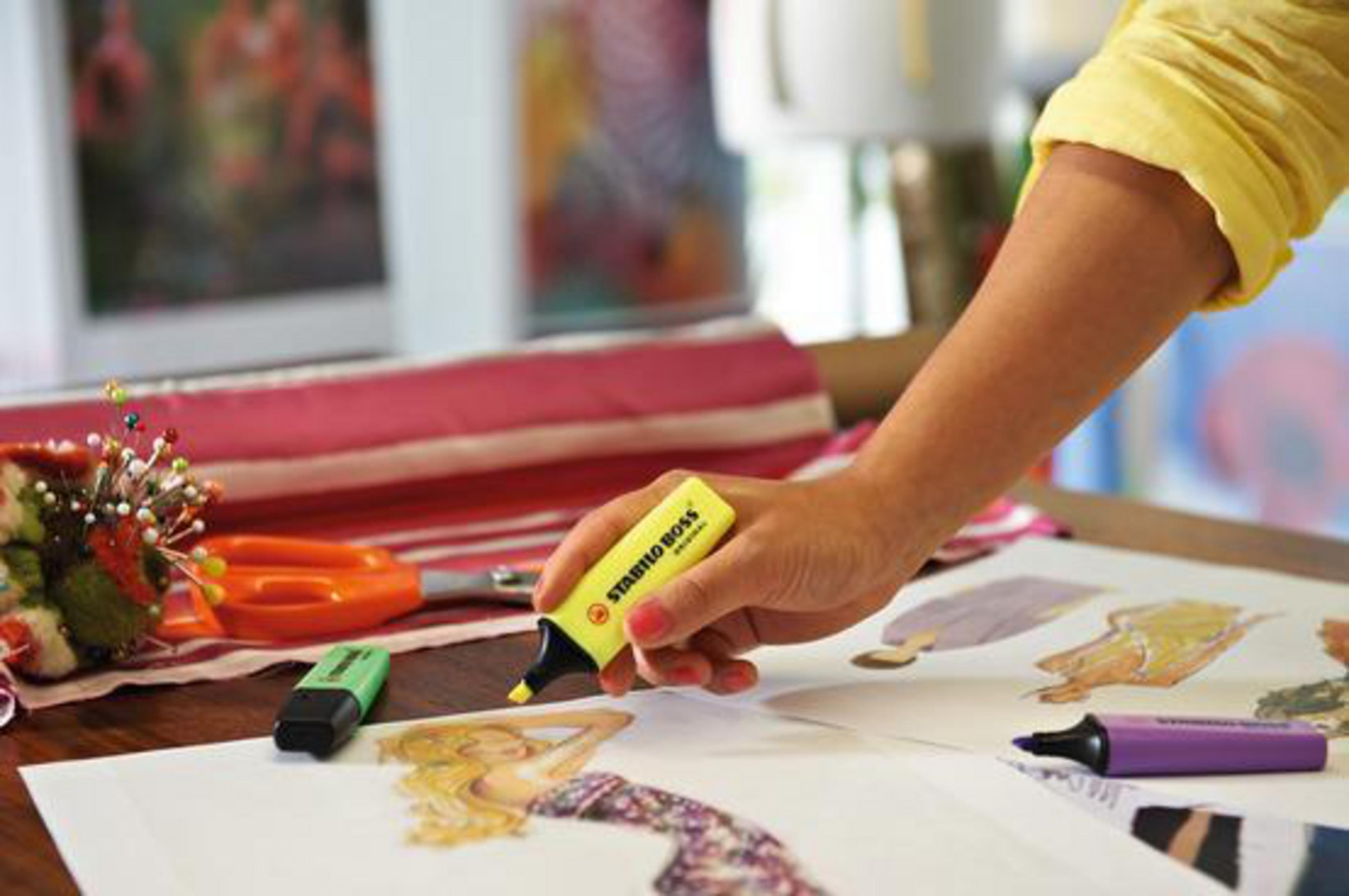 Una mano tiene un evidenziatore giallo su schizzi di moda. Forbici e un evidenziatore verde nelle vicinanze.