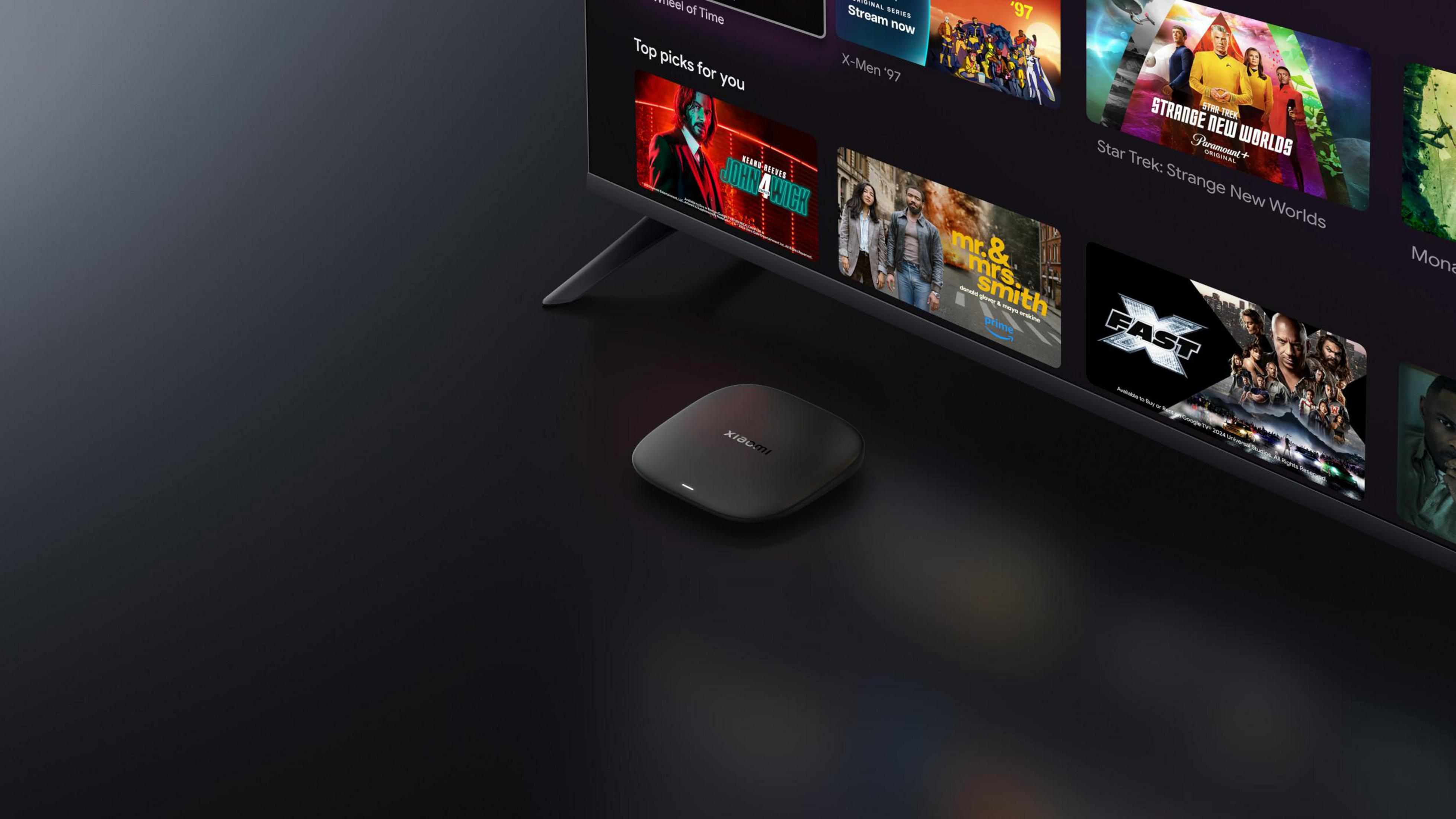 Bir Xiaomi TV yayın cihazı, film seçenekleri gösteren modern bir TV'nin yanında duruyor.