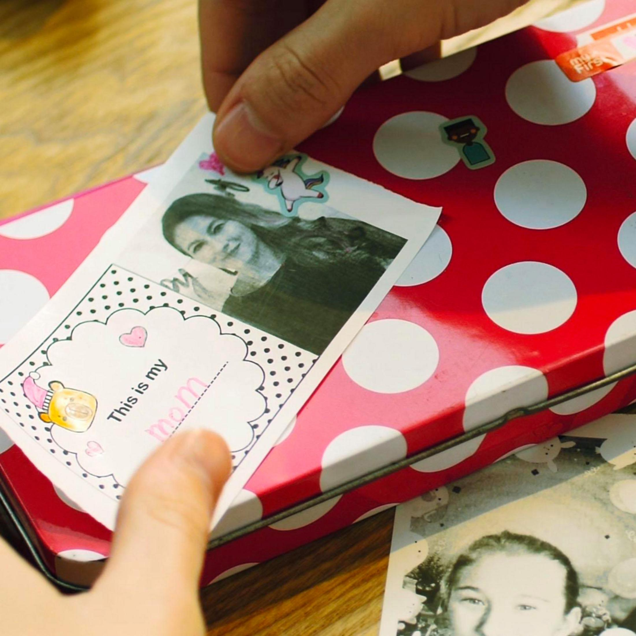 Las manos colocan una tarjeta con foto de mamá sobre una caja de lunares rojos y blancos.
