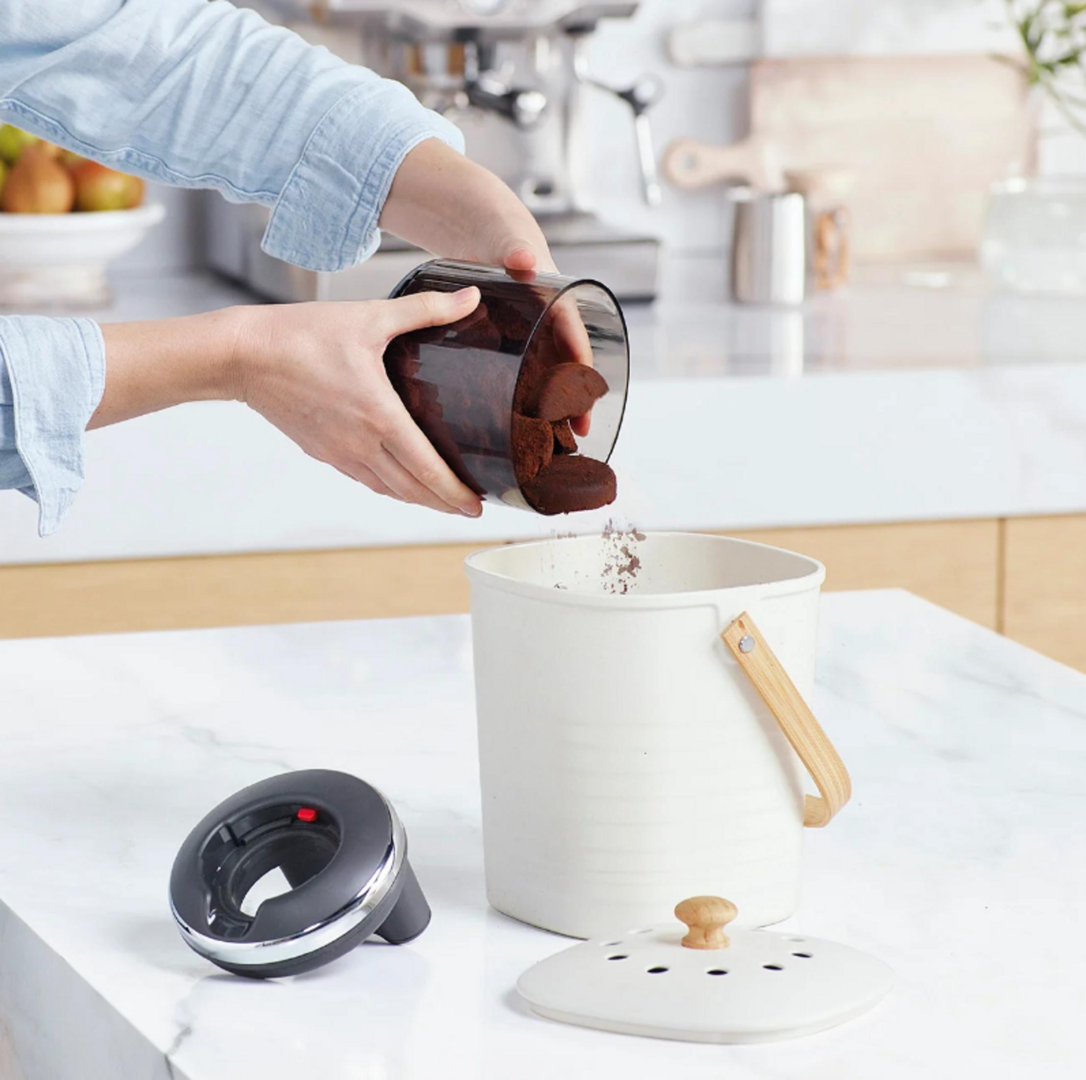 Una persona versa fondi di caffè da un contenitore trasparente in un bidone bianco.
