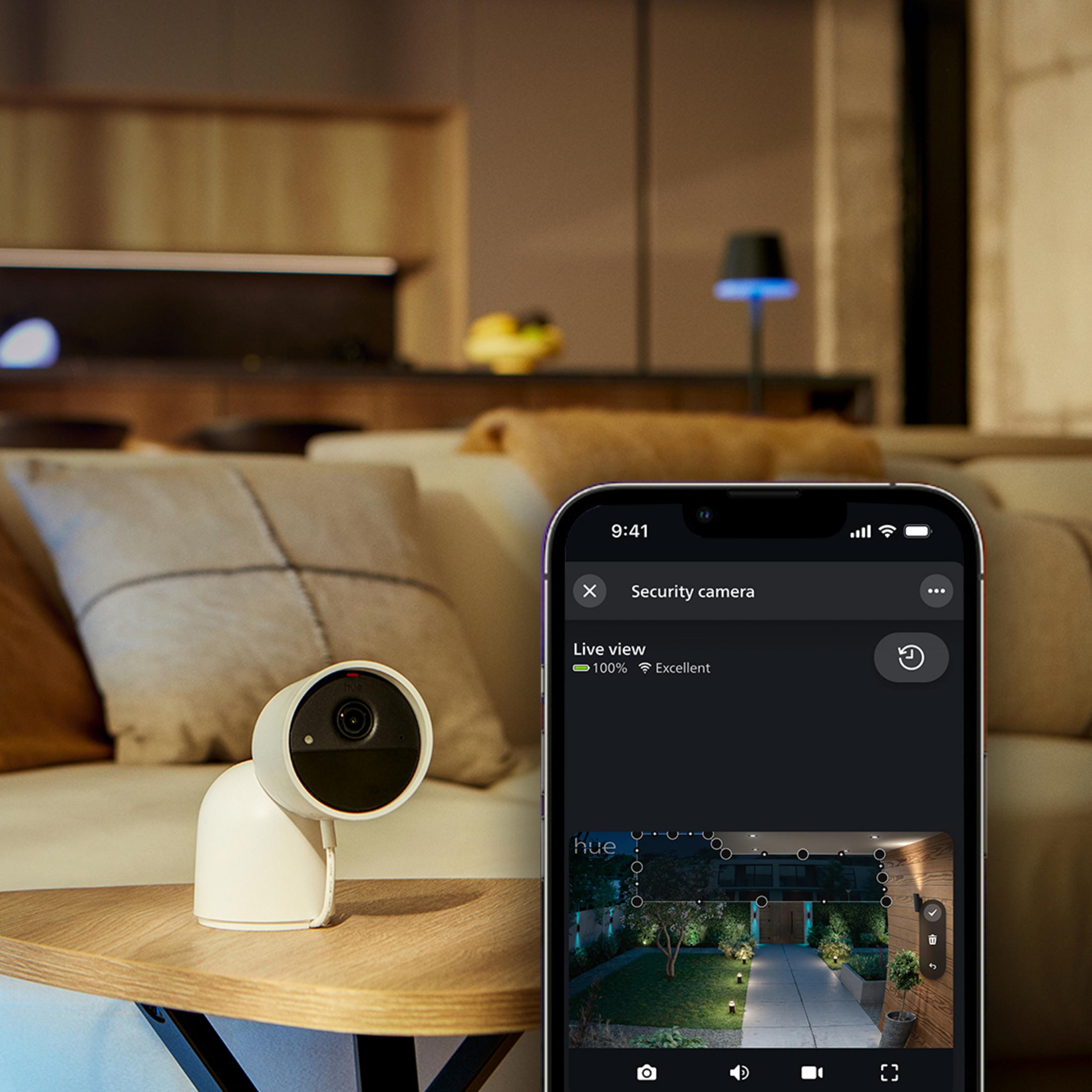 Eine weiße Überwachungskamera neben einem Smartphone, das eine Gartenansicht zeigt.