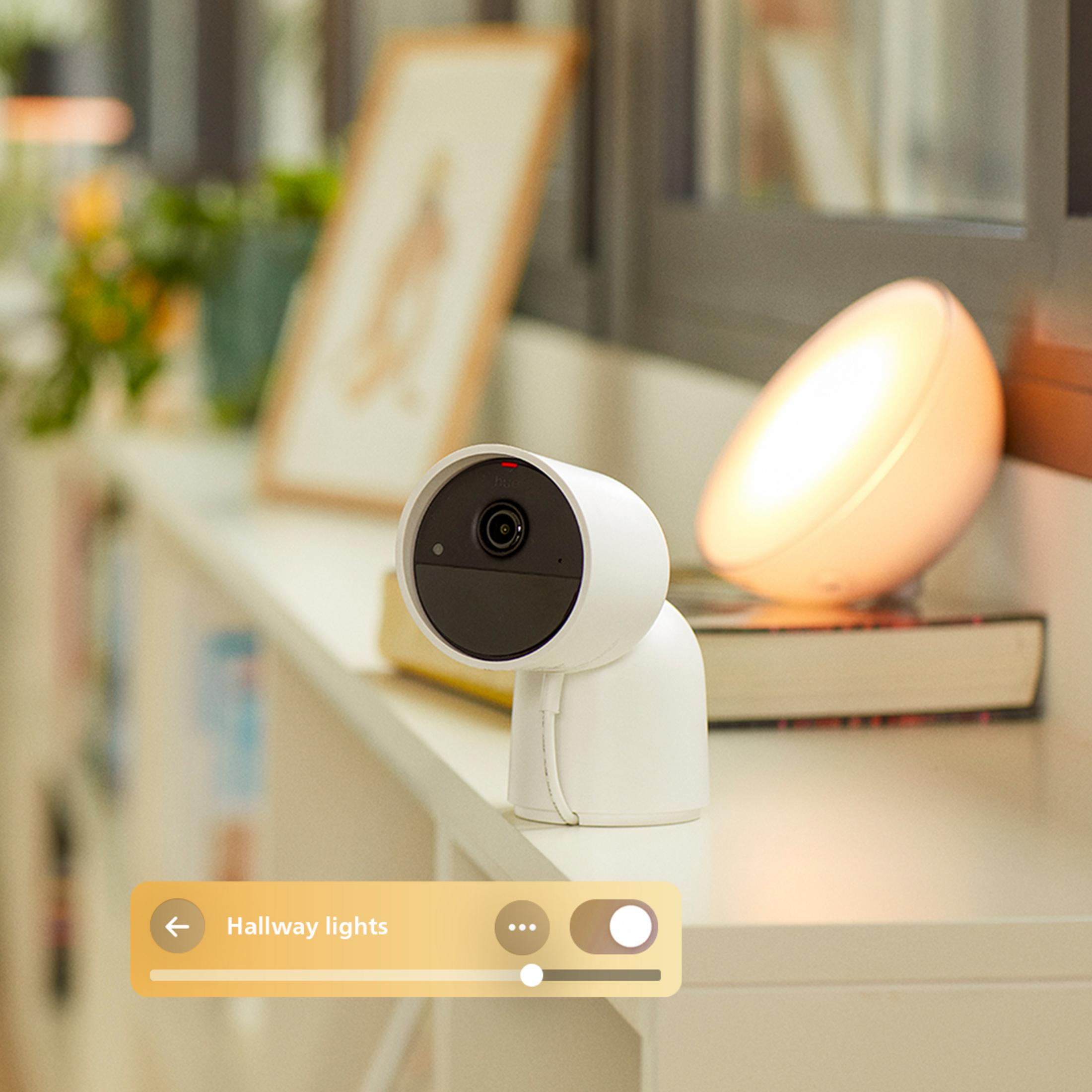 Ein weißer Smart-Kamerakopf mit rotem Licht auf einem Regal, daneben ein Lampenlicht.