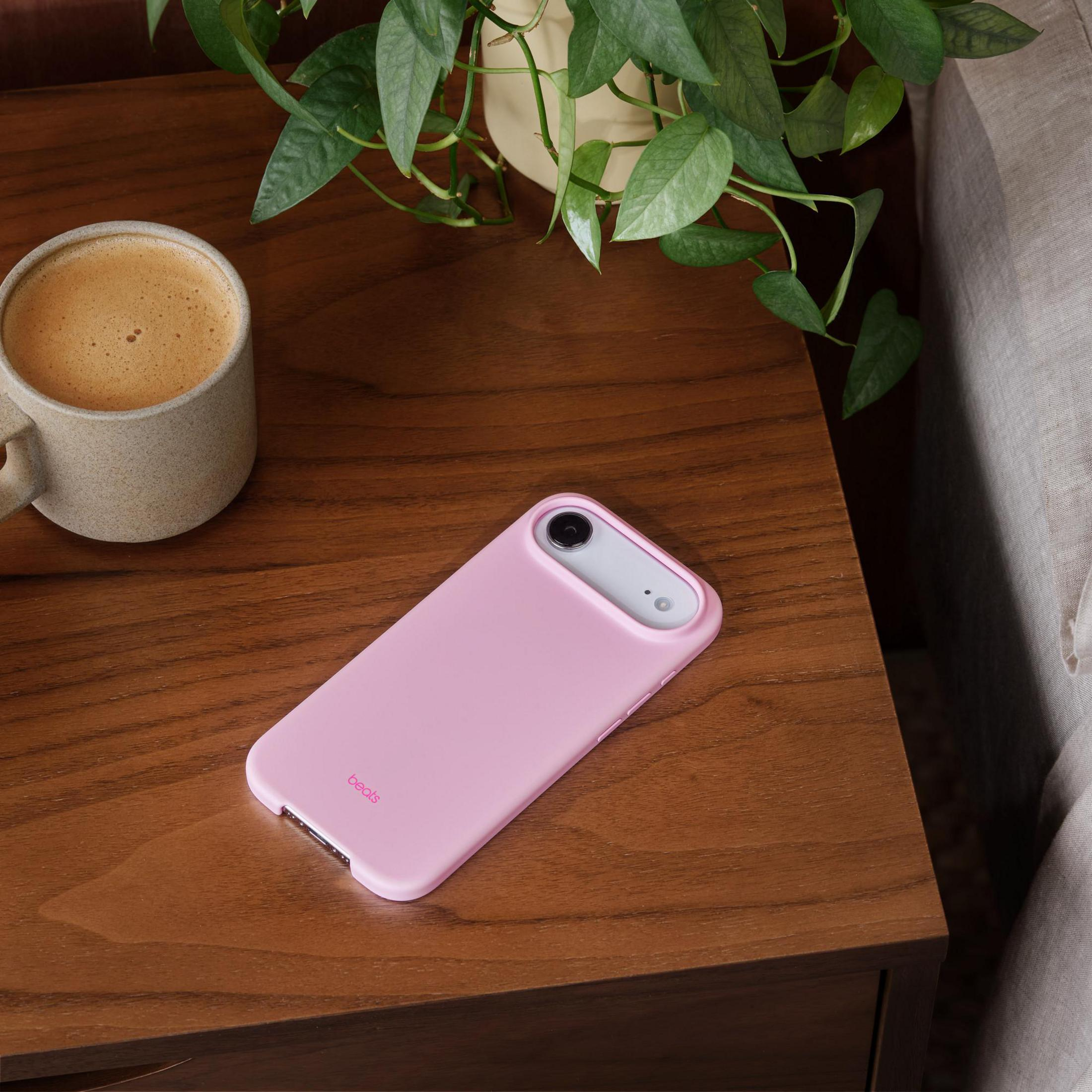 Pink phone case with Beats logo on wooden table next to coffee and plant.