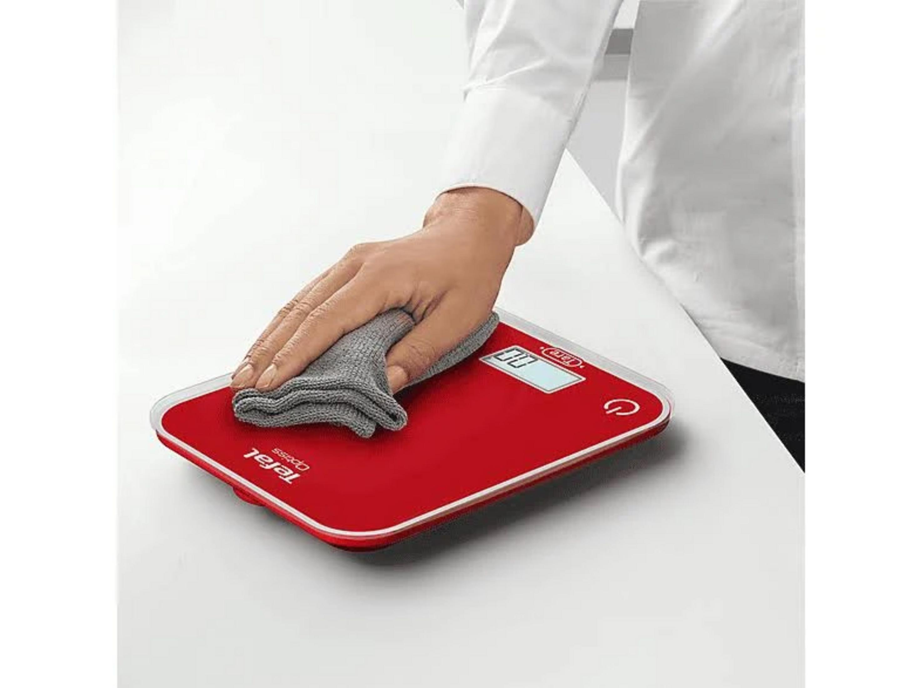 Eine Hand reinigt eine rote Küchenwaage mit einem grauen Tuch.