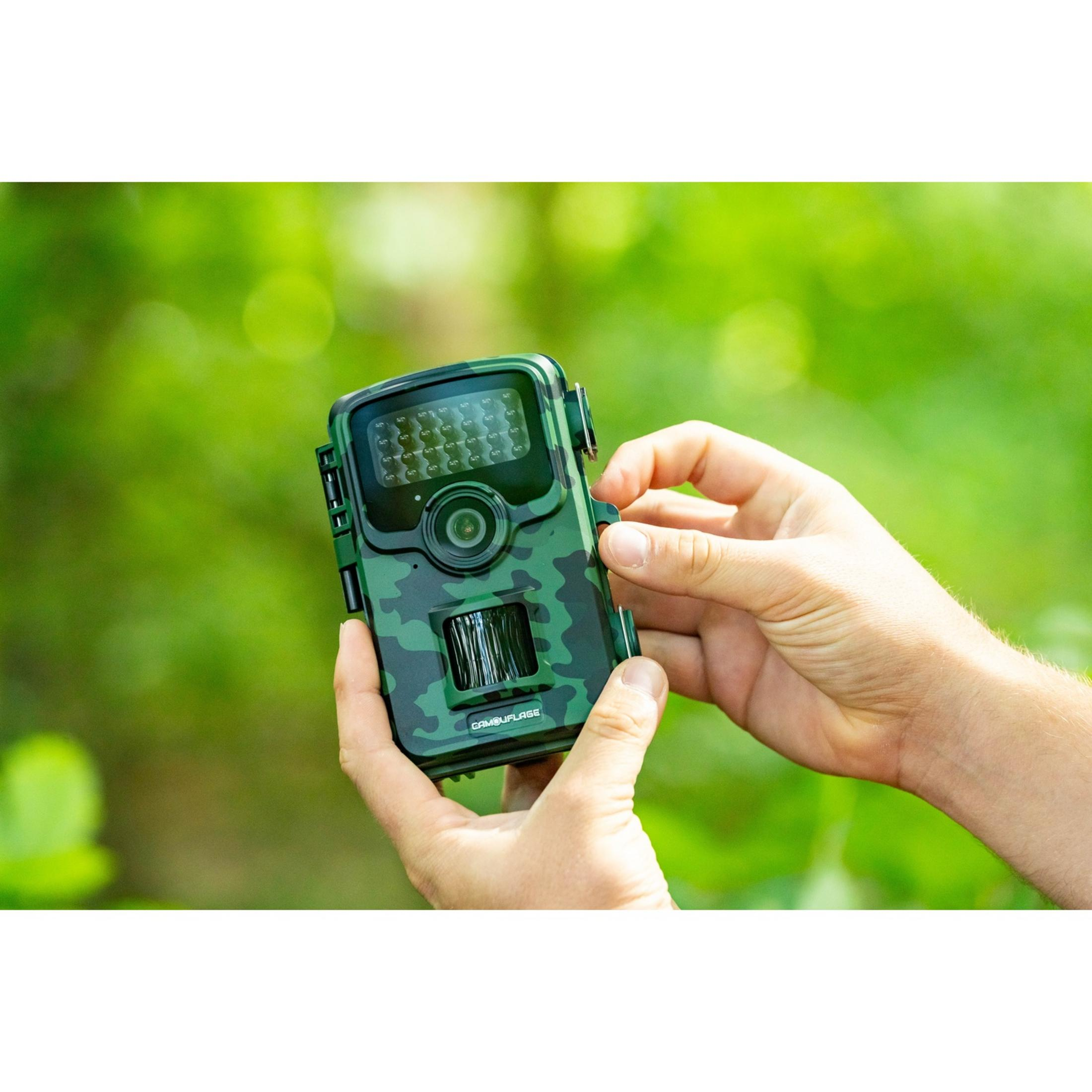 Una mano tiene una telecamera mimetica verde su uno sfondo verde sfocato.