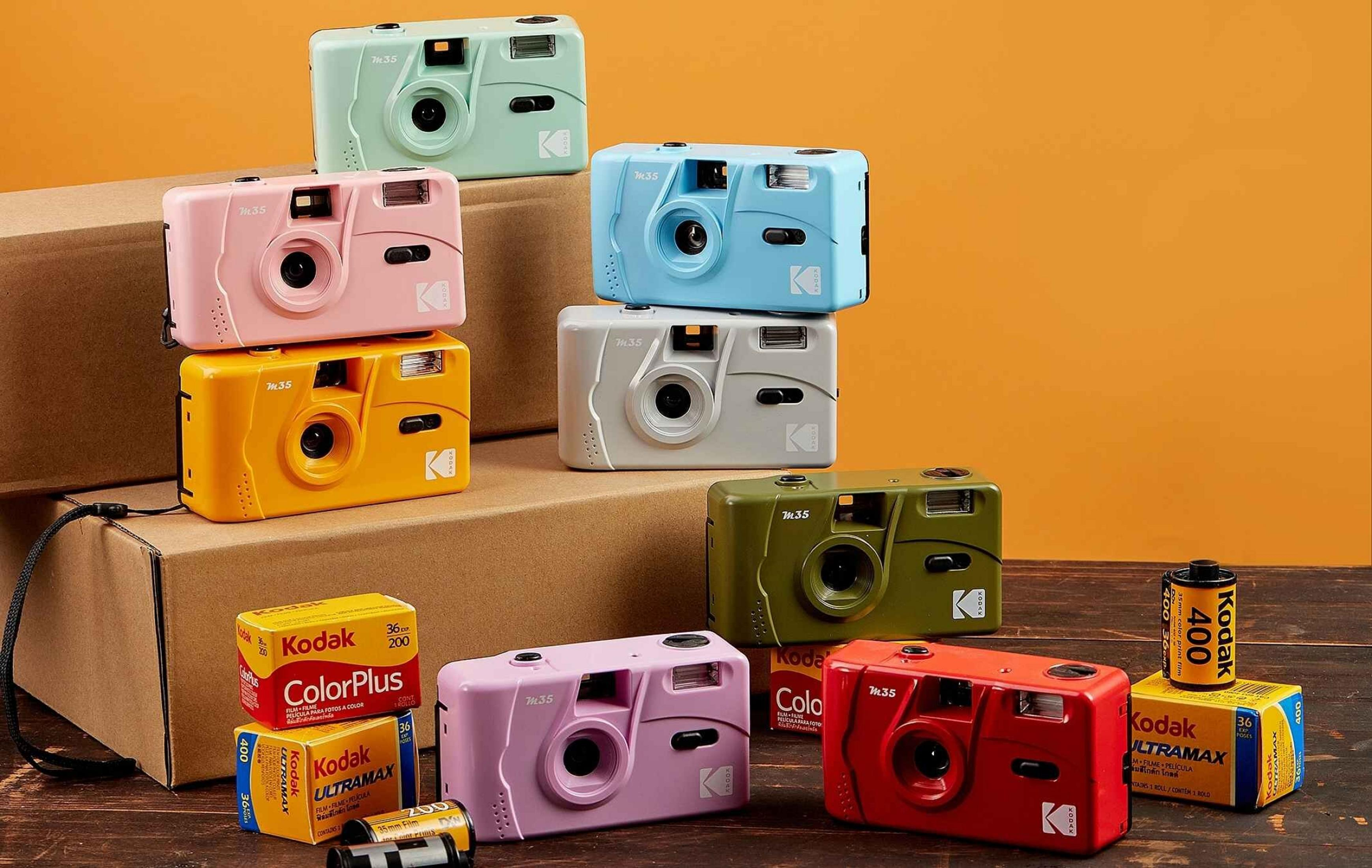 Diverse fotocamere Kodak M35 colorate e rullini sono disposti su scatole di cartone e un tavolo di legno.