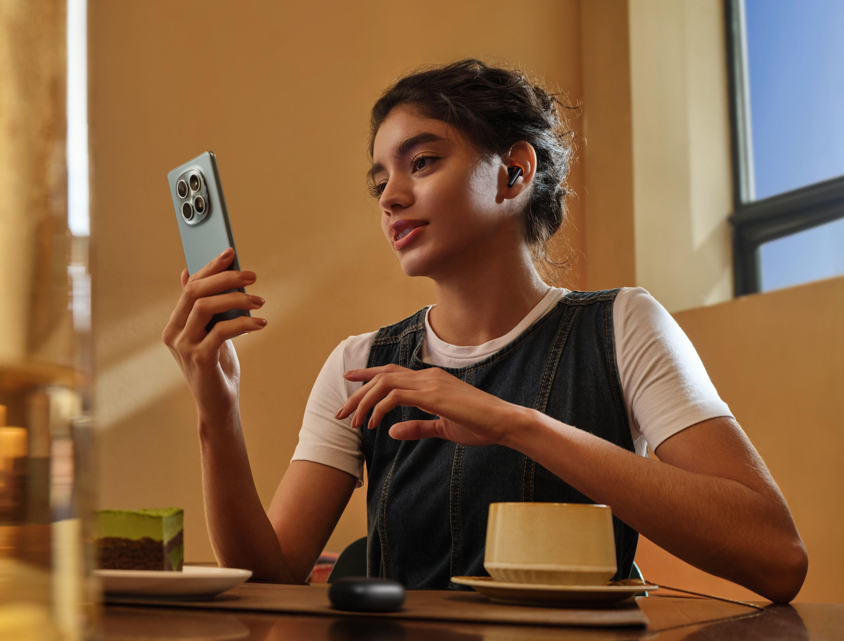 Una donna con auricolari tiene un telefono; torta e tazza sul tavolo.