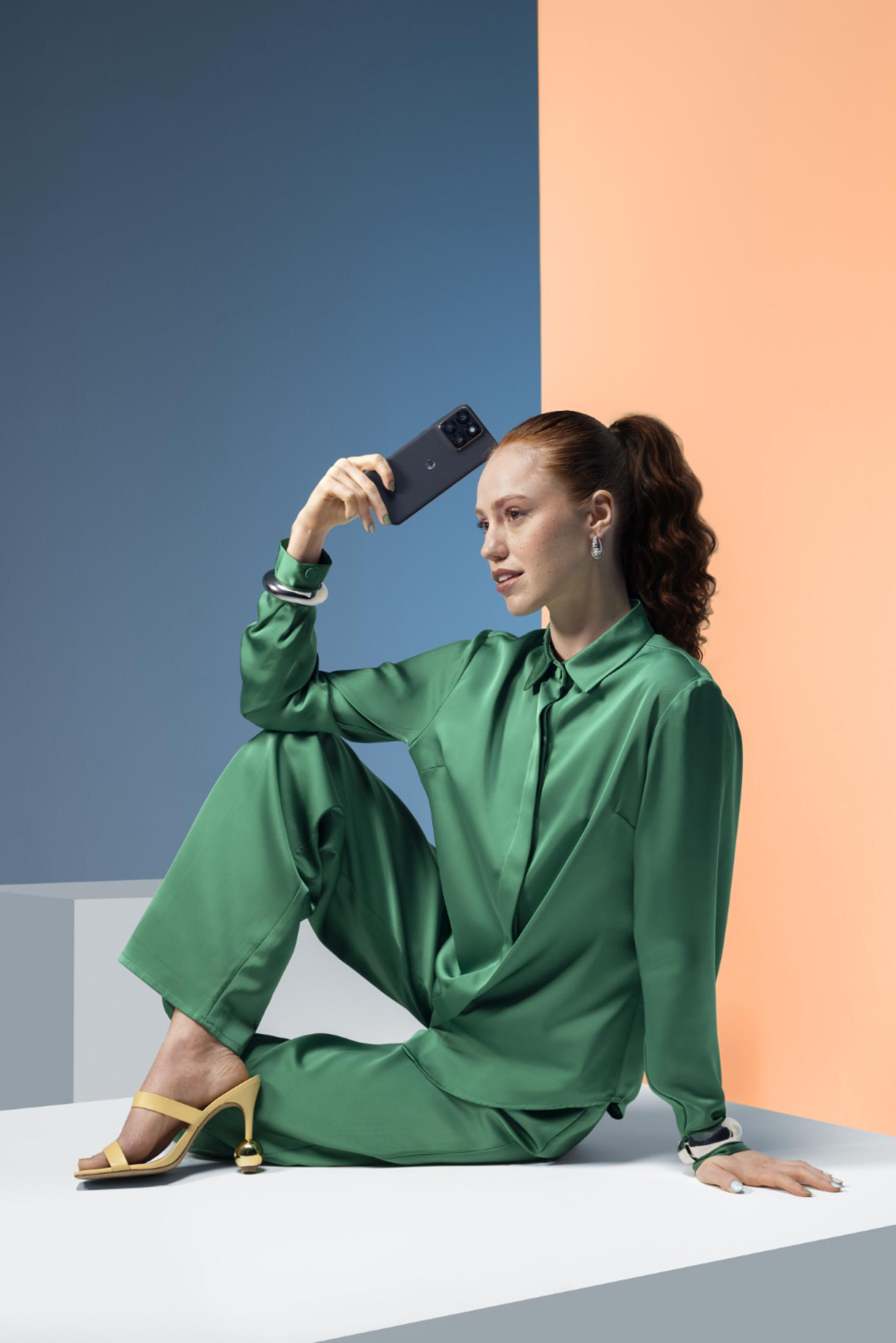 Una donna in un completo di raso verde è seduta tenendo uno smartphone sopra la testa.