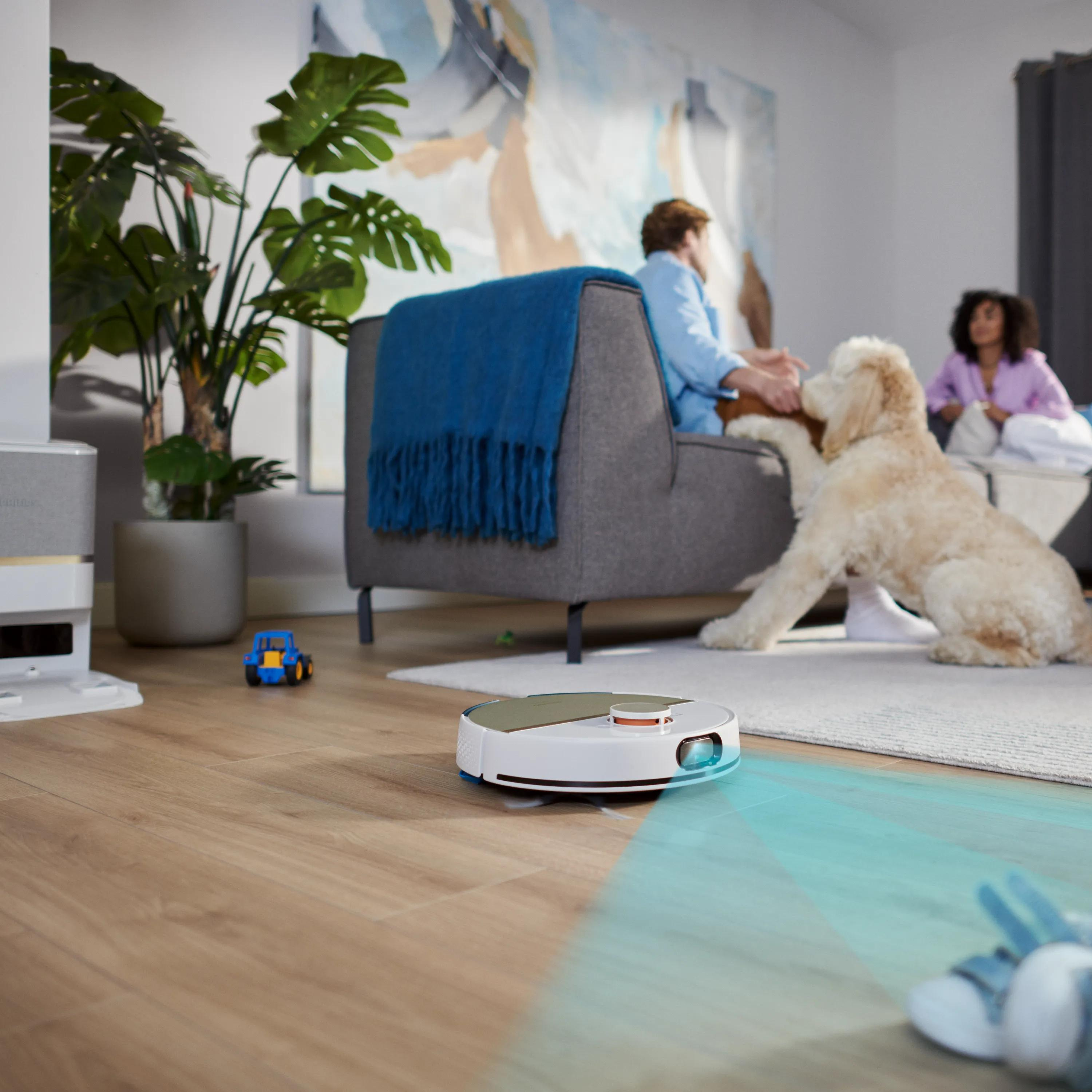 Un robot aspirapolvere pulisce un pavimento in legno mentre una famiglia e un cane sono sullo sfondo.