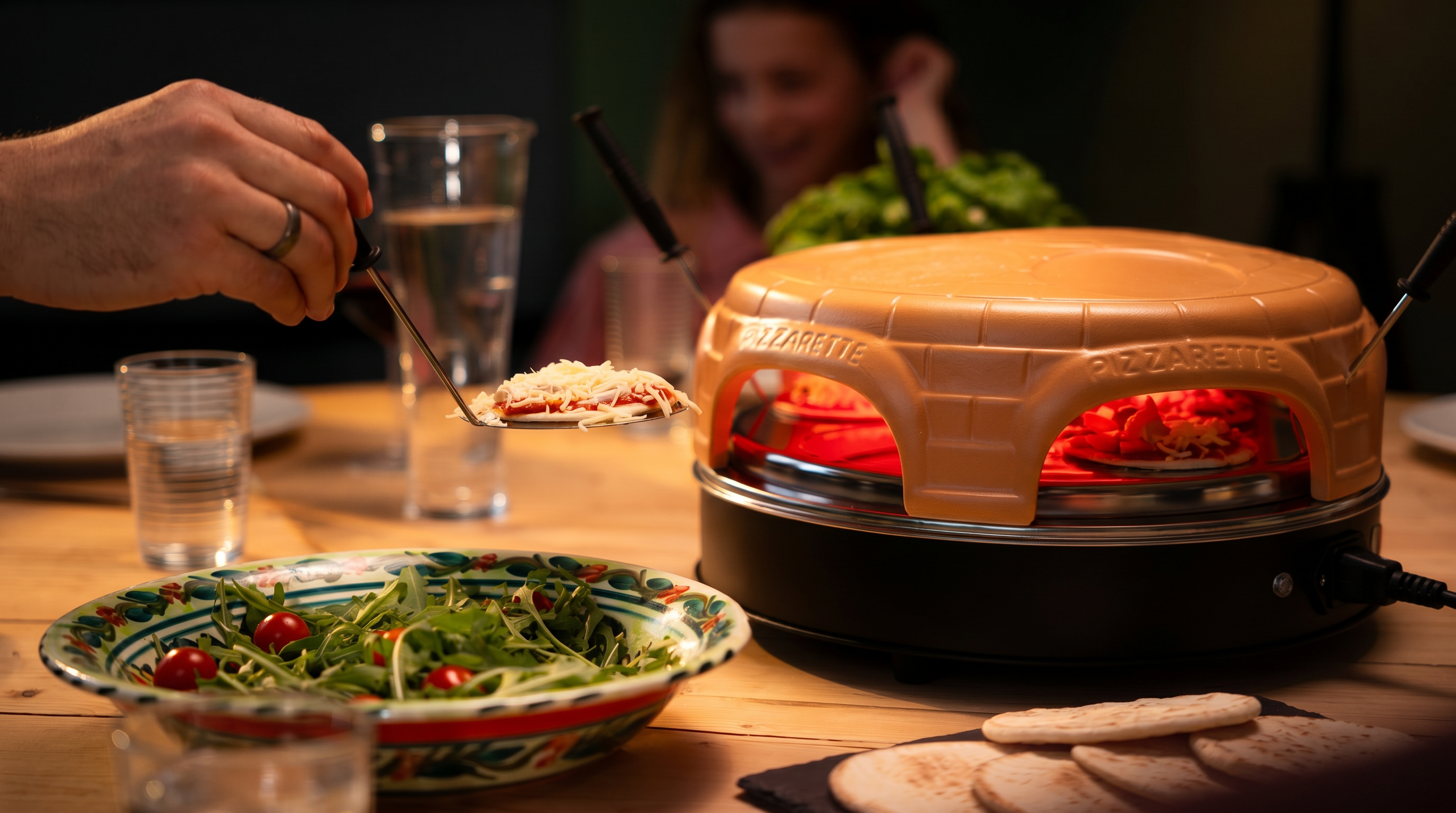 Una mano posiziona una mini pizza su un forno pizzarette, con un'insalata vicina.