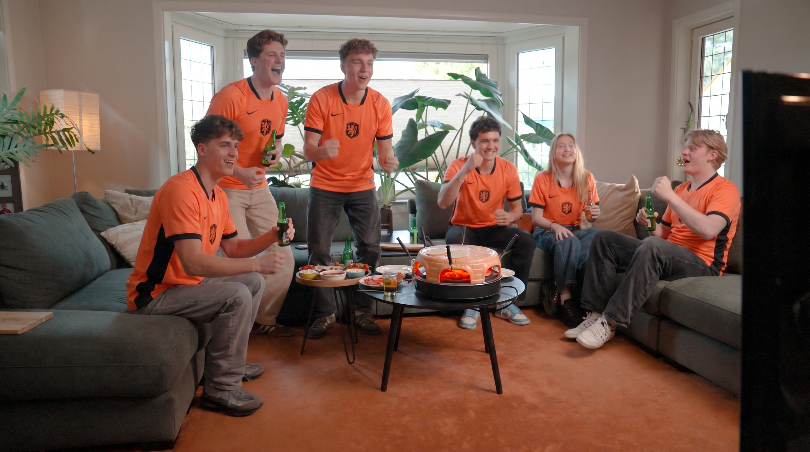 Zes jongeren in oranje shirts juichen voor een tv, met snacks en drankjes op tafel.