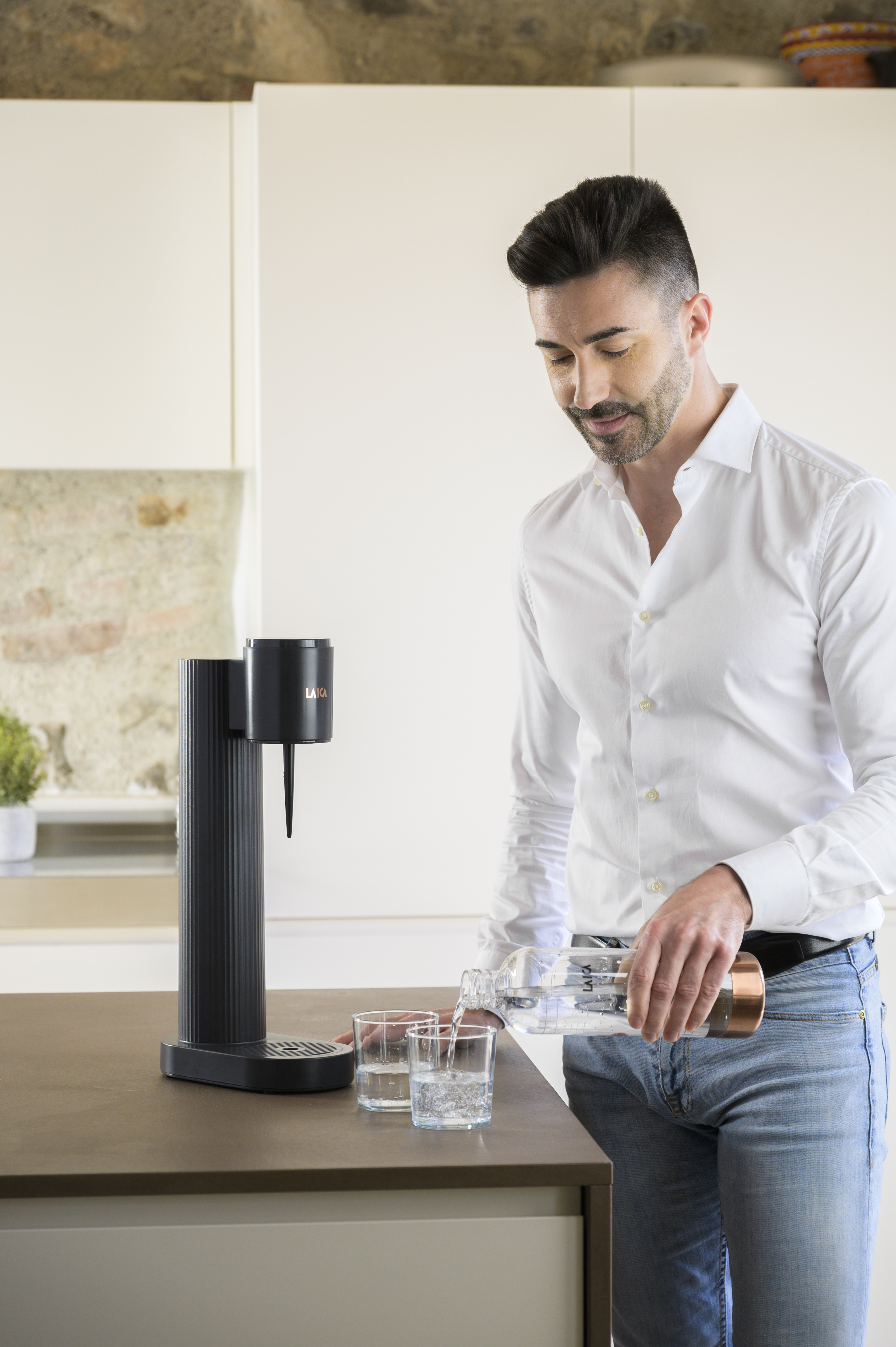Hombre con camisa blanca y jeans vierte agua de botella en vasos junto a dispensador.