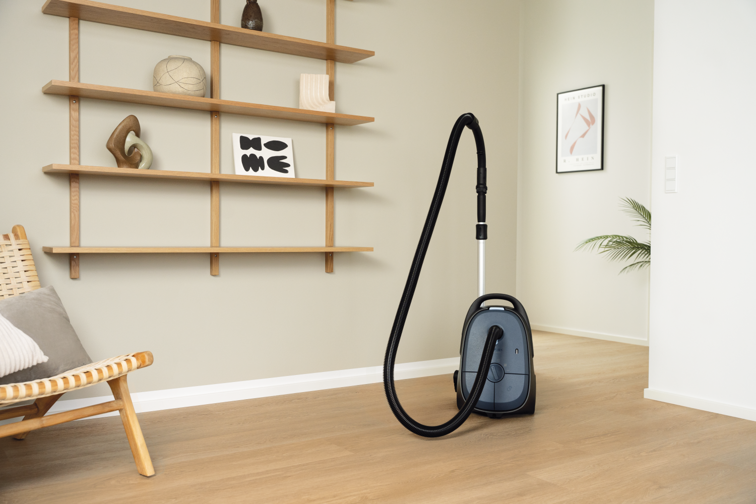 Een grijze stofzuiger staat op een houten vloer naast een stoel. Planken zijn aan de muur.