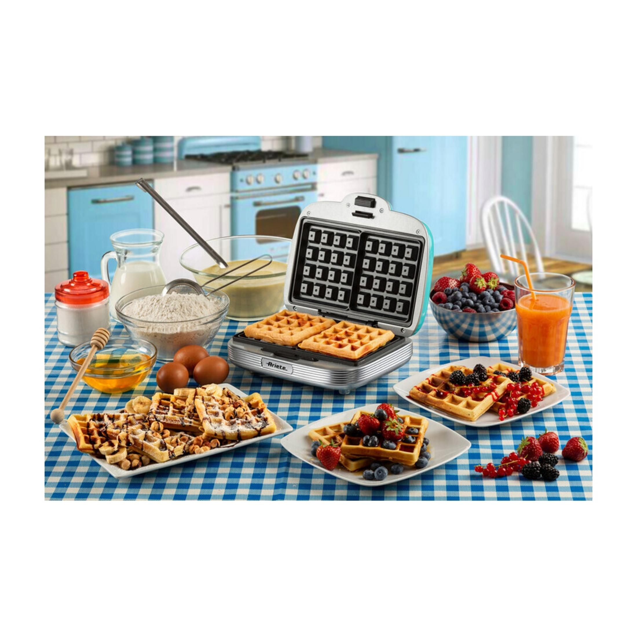 Waffeleisen mit fertigen Waffeln, Obst und Zutaten auf karierter Tischdecke.