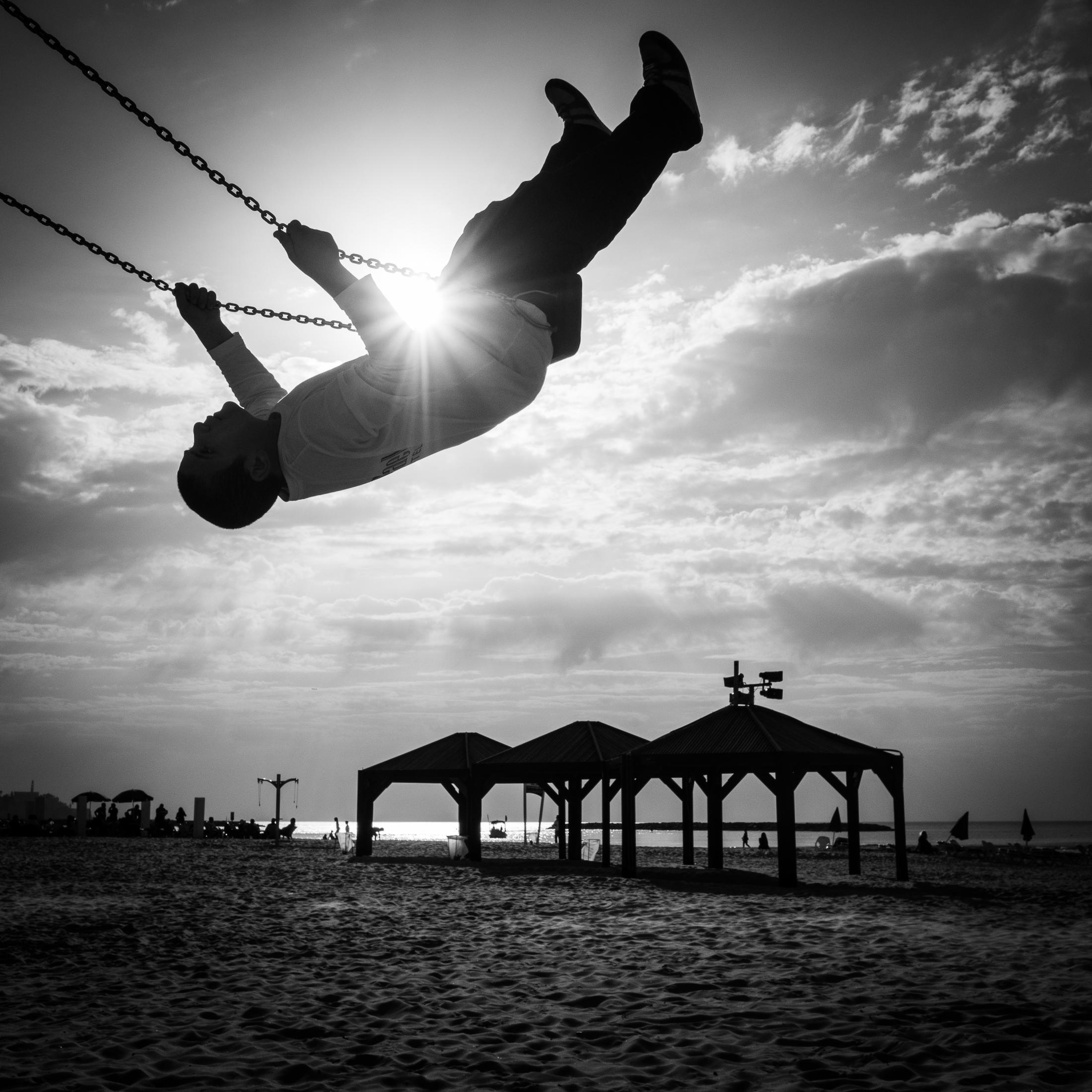 Kind schaukelt hoch gegen die Sonne, Strand und Meer im Hintergrund.