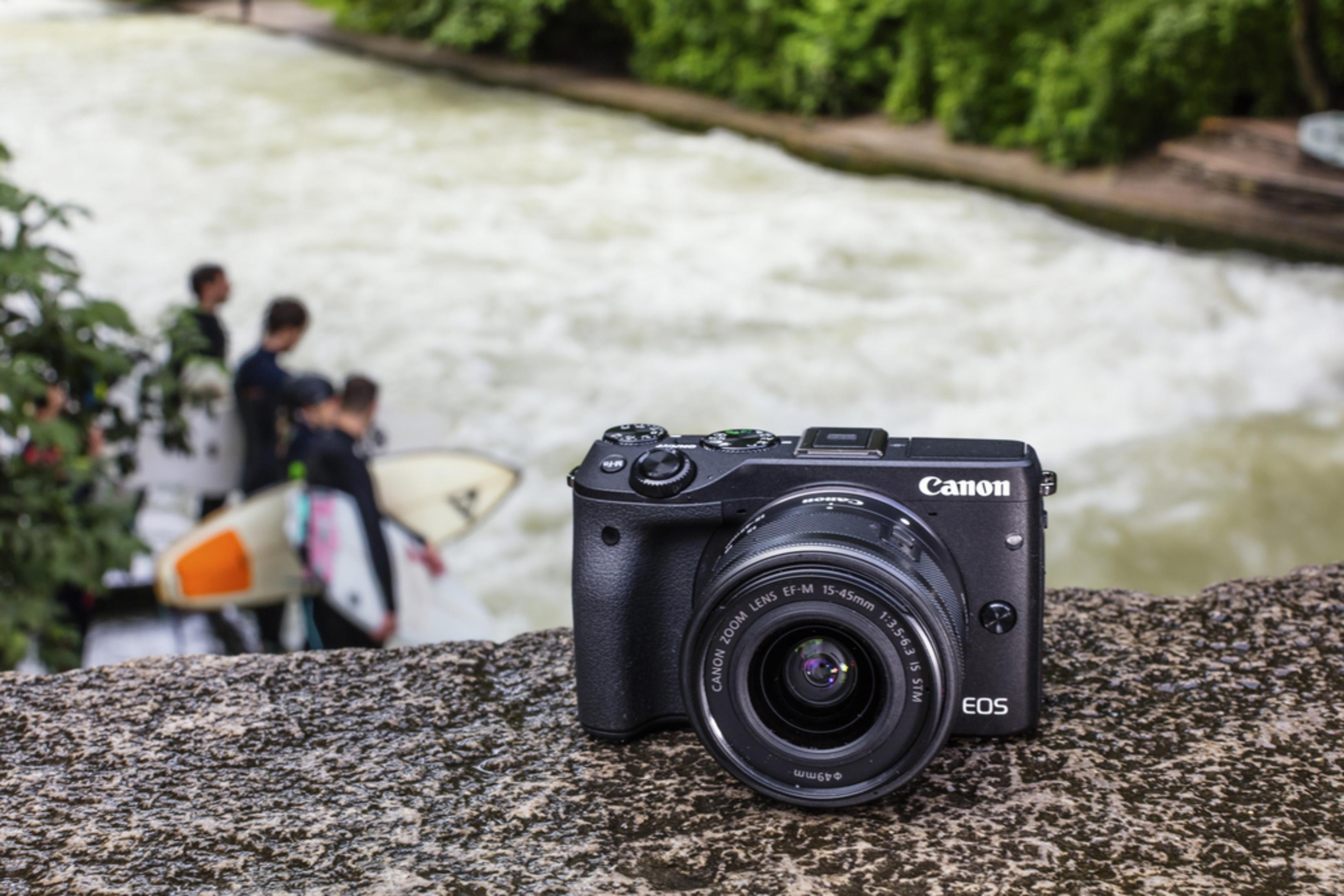 Eine schwarze Canon EOS Kamera liegt auf einem Stein. Im Hintergrund surfen Menschen in einem Fluss.