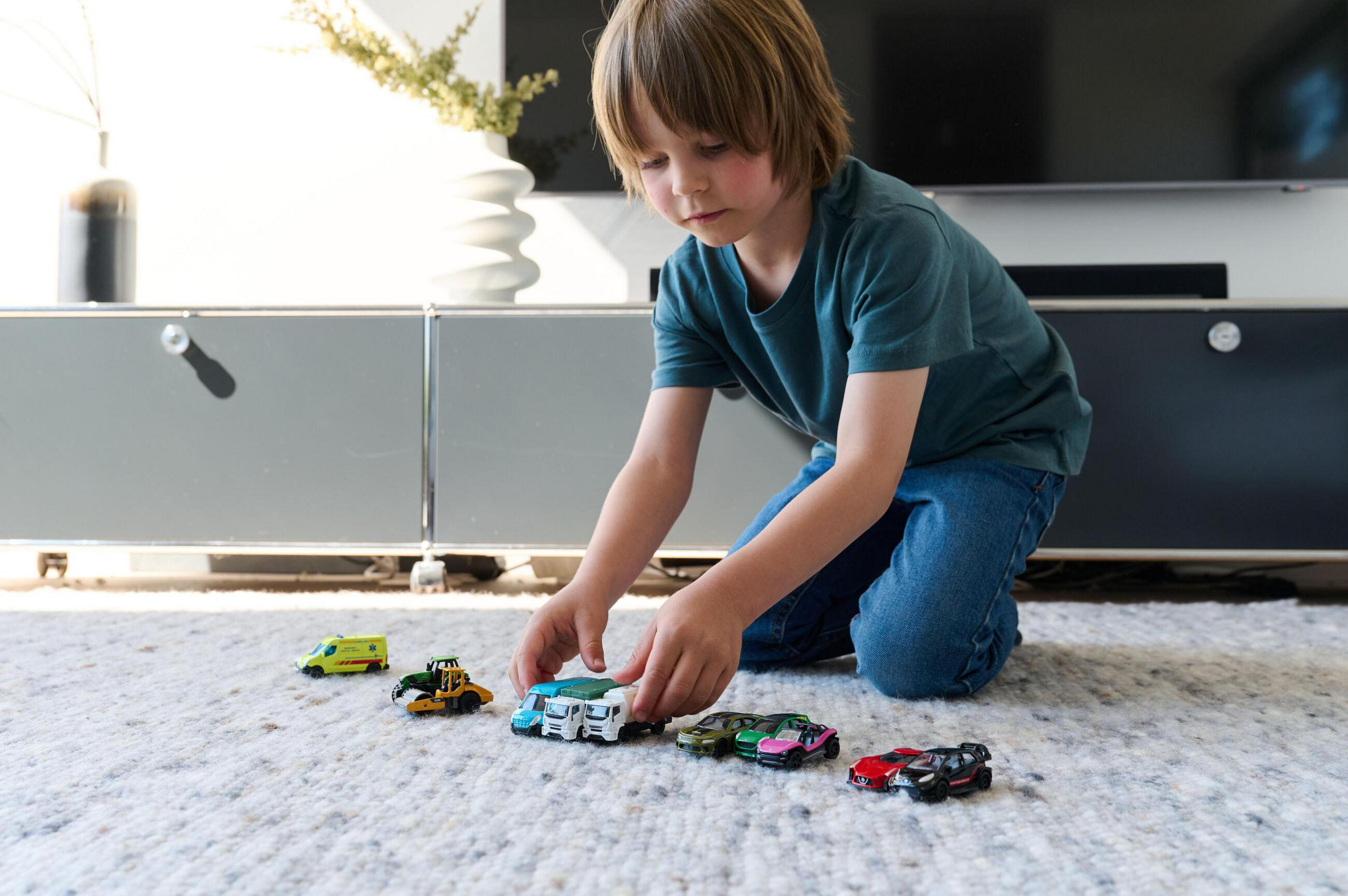 Ein Junge spielt mit Spielzeugautos auf einem Teppich. Zu sehen sind ein Krankenwagen, ein Bagger und mehrere Autos.