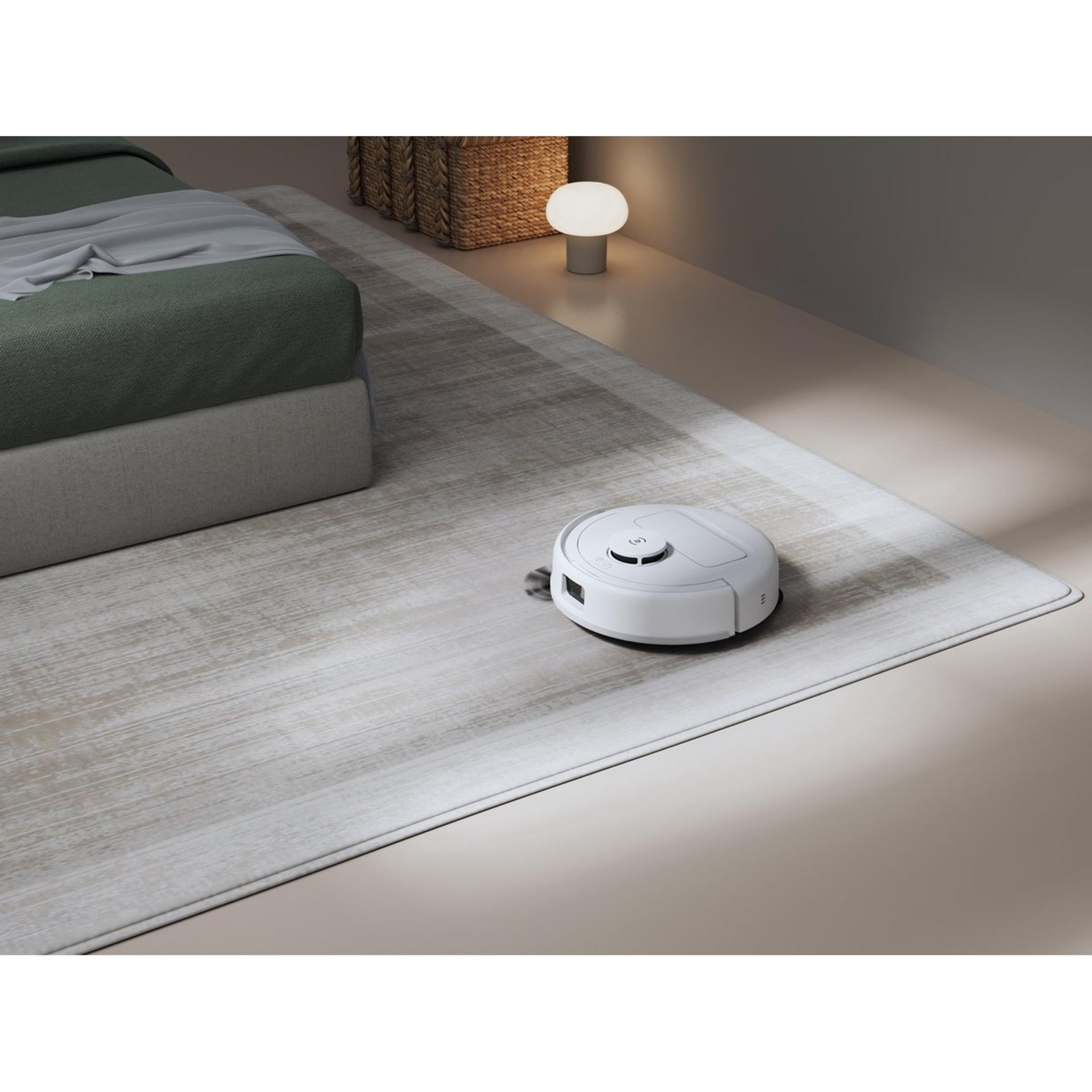 Ein weißer Saugroboter fährt auf einem hellen Teppich neben einem Bett.