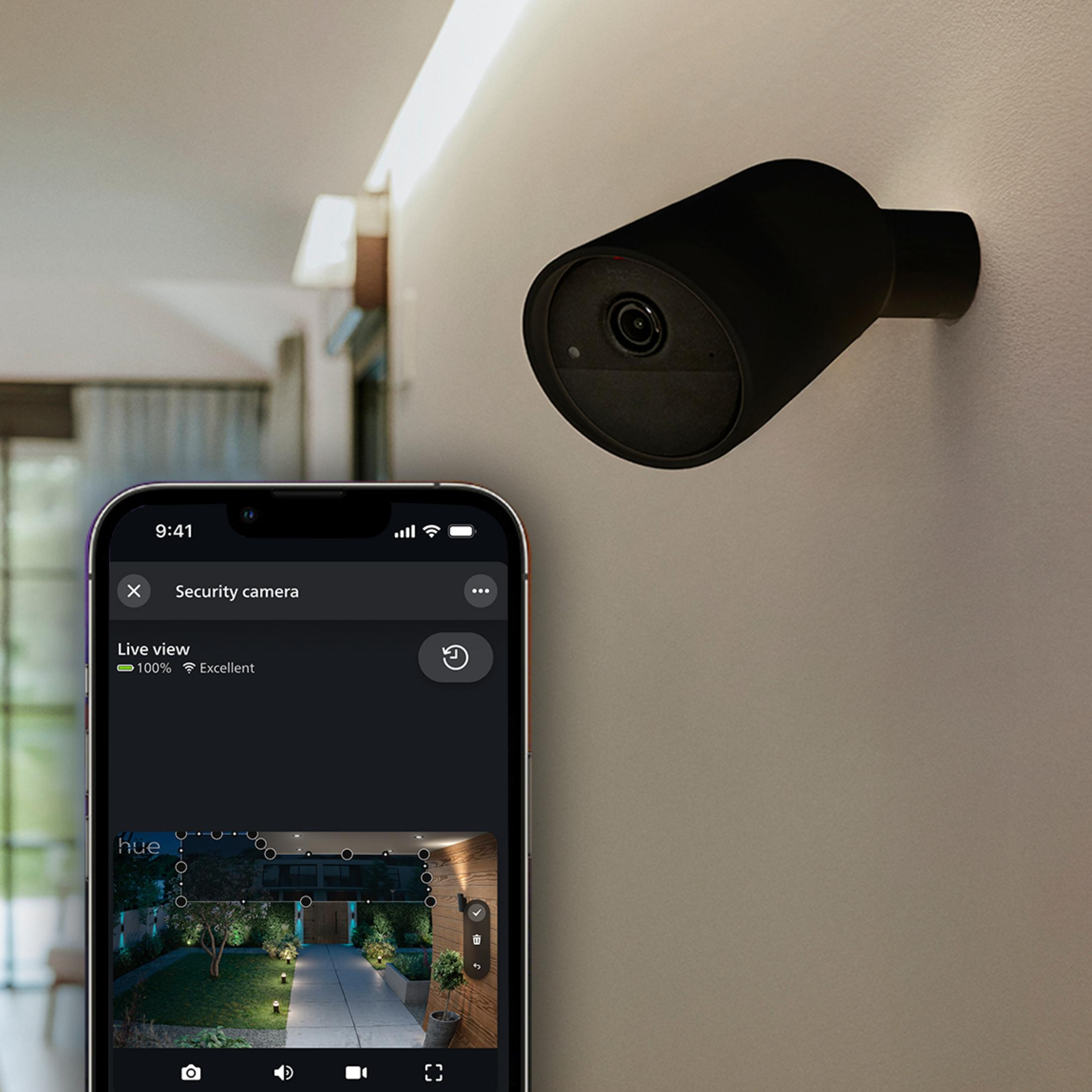 Una telecamera di sicurezza nera su un muro, accanto a uno smartphone che mostra una vista dal vivo.