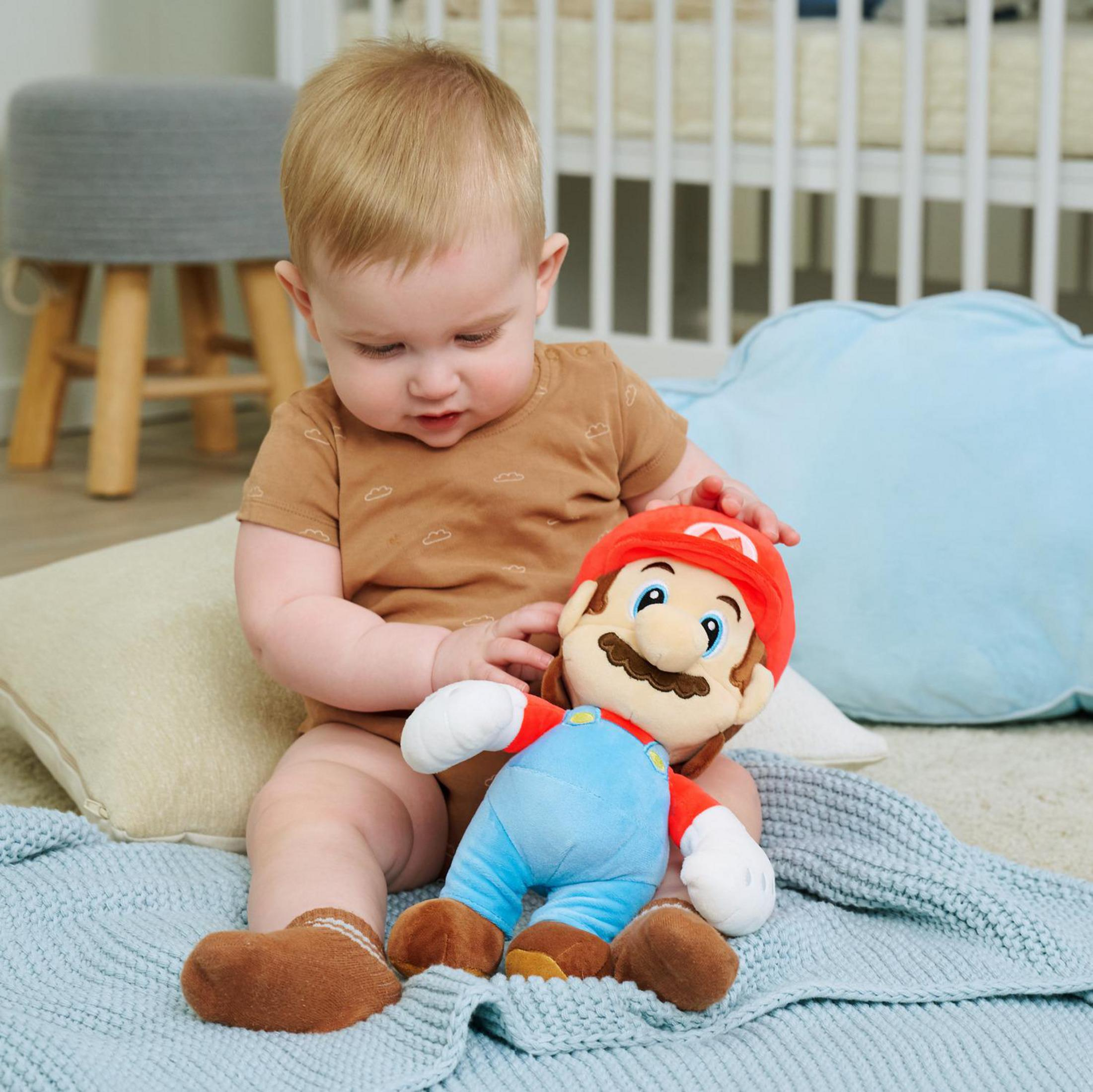 Baby in brauner Kleidung hält Mario-Plüschtier auf blauer Decke nahe Kinderbett.