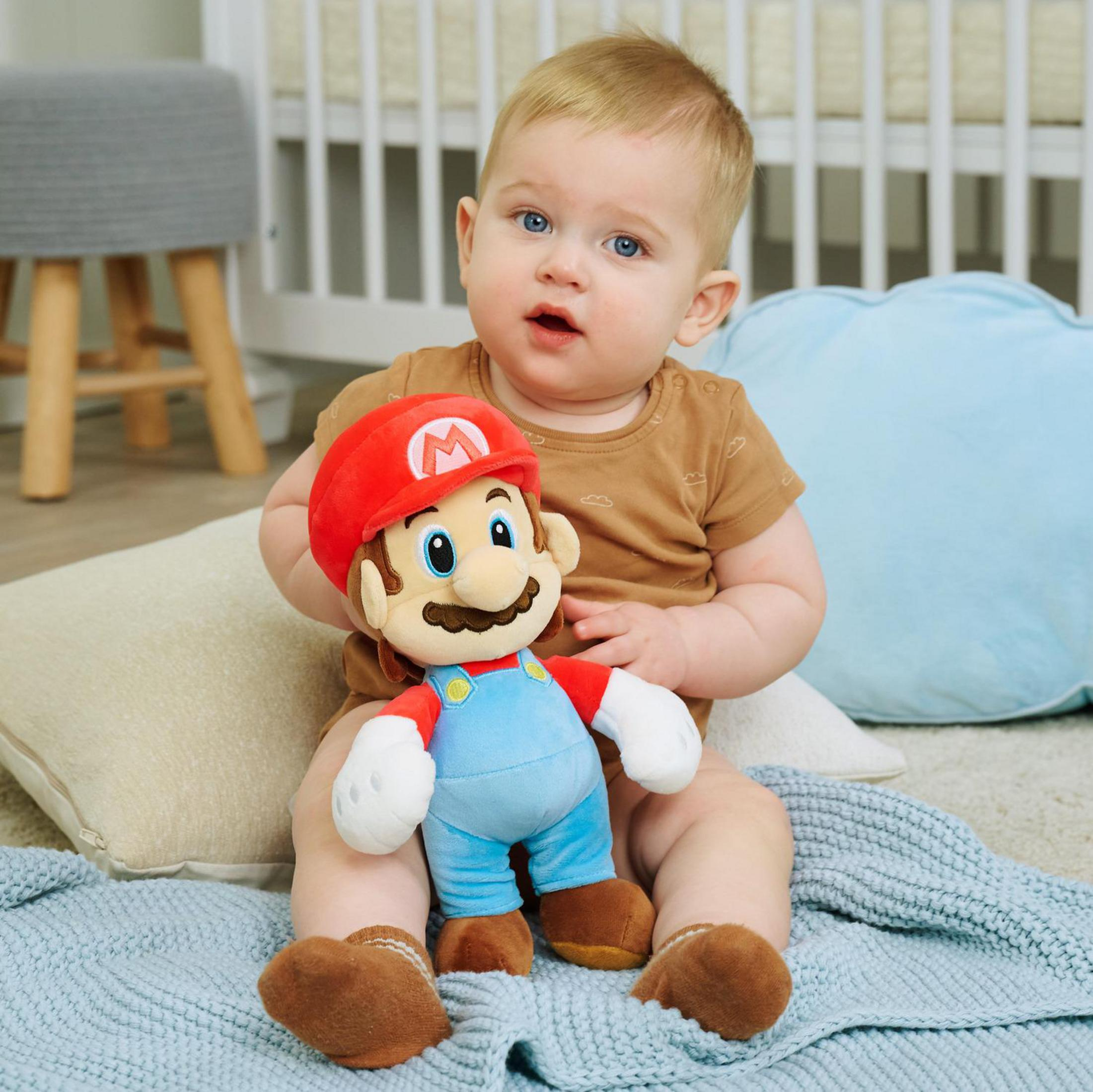 Ein Baby sitzt und hält eine Mario-Puppe. Ein Kinderbett ist im Hintergrund.