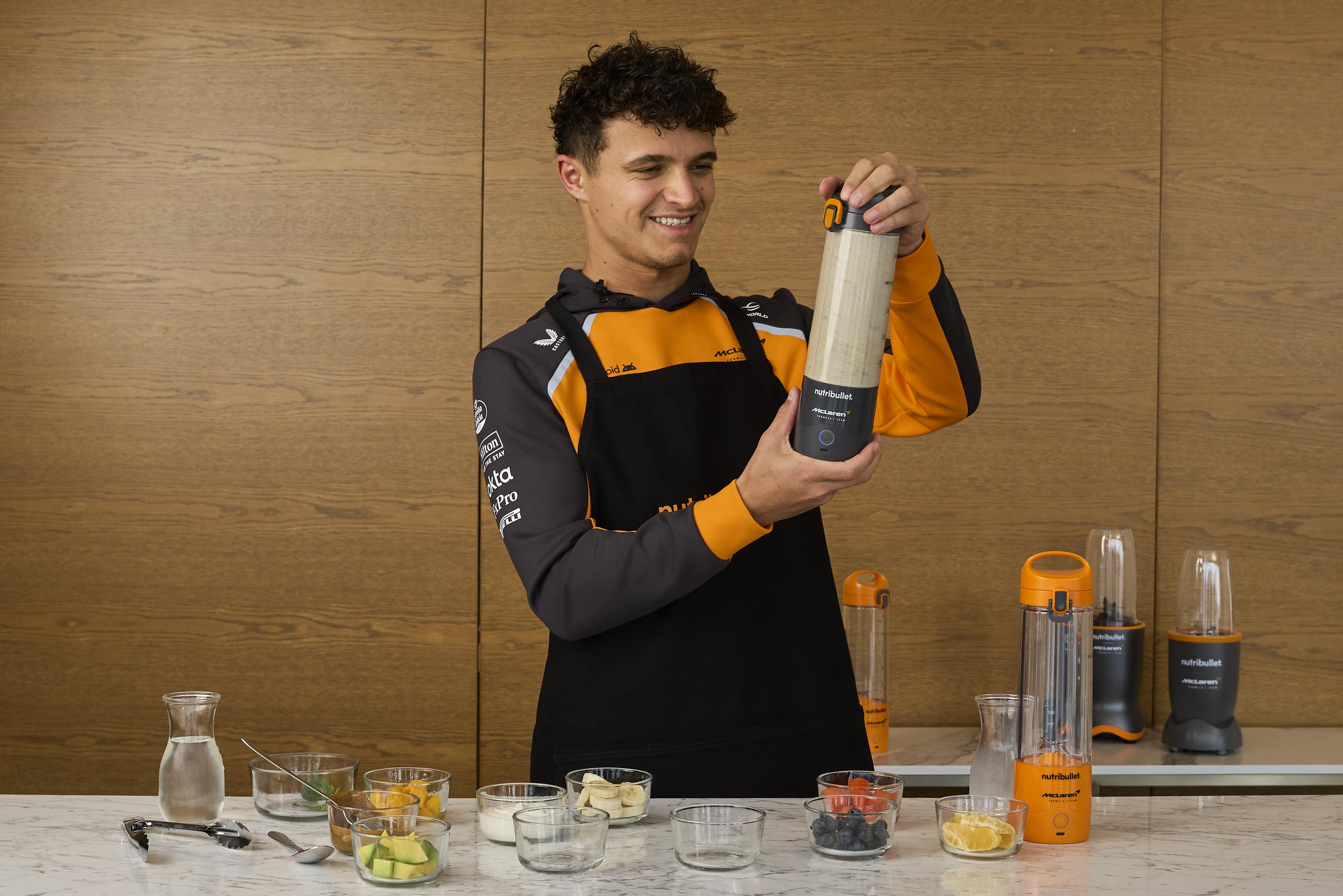 Ein Mann in einem McLaren-Outfit hält einen Nutribullet-Becher vor einer Holzwand.