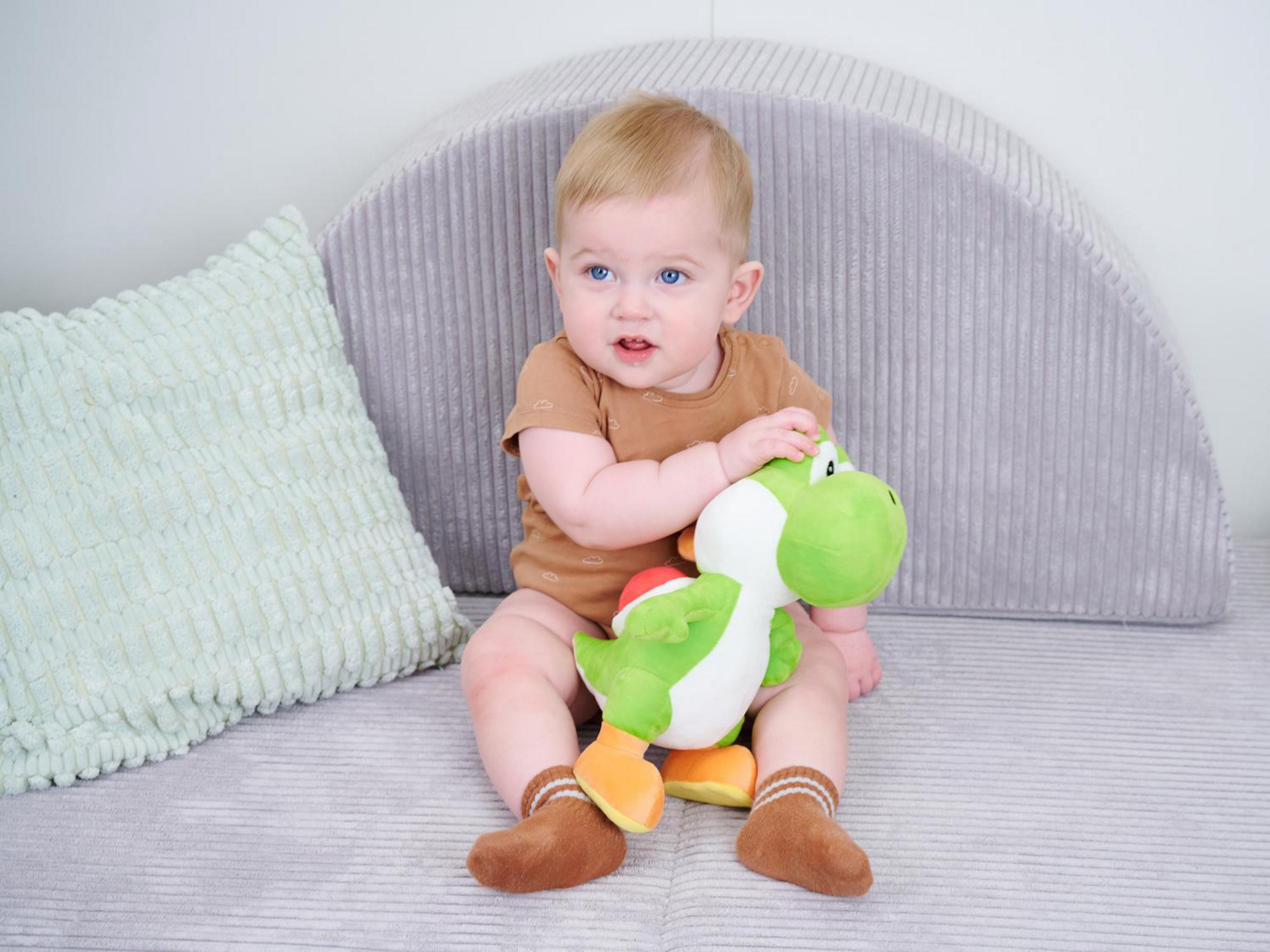 Ein Baby sitzt auf einem Sofa und hält eine grüne Plüschfigur.