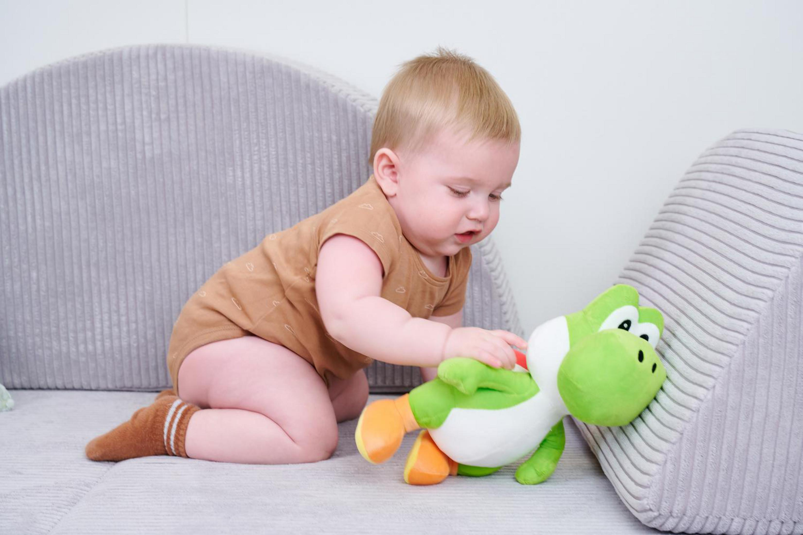 Ein Baby in brauner Kleidung spielt mit einem grünen Yoshi-Plüschtier auf einem Sofa.