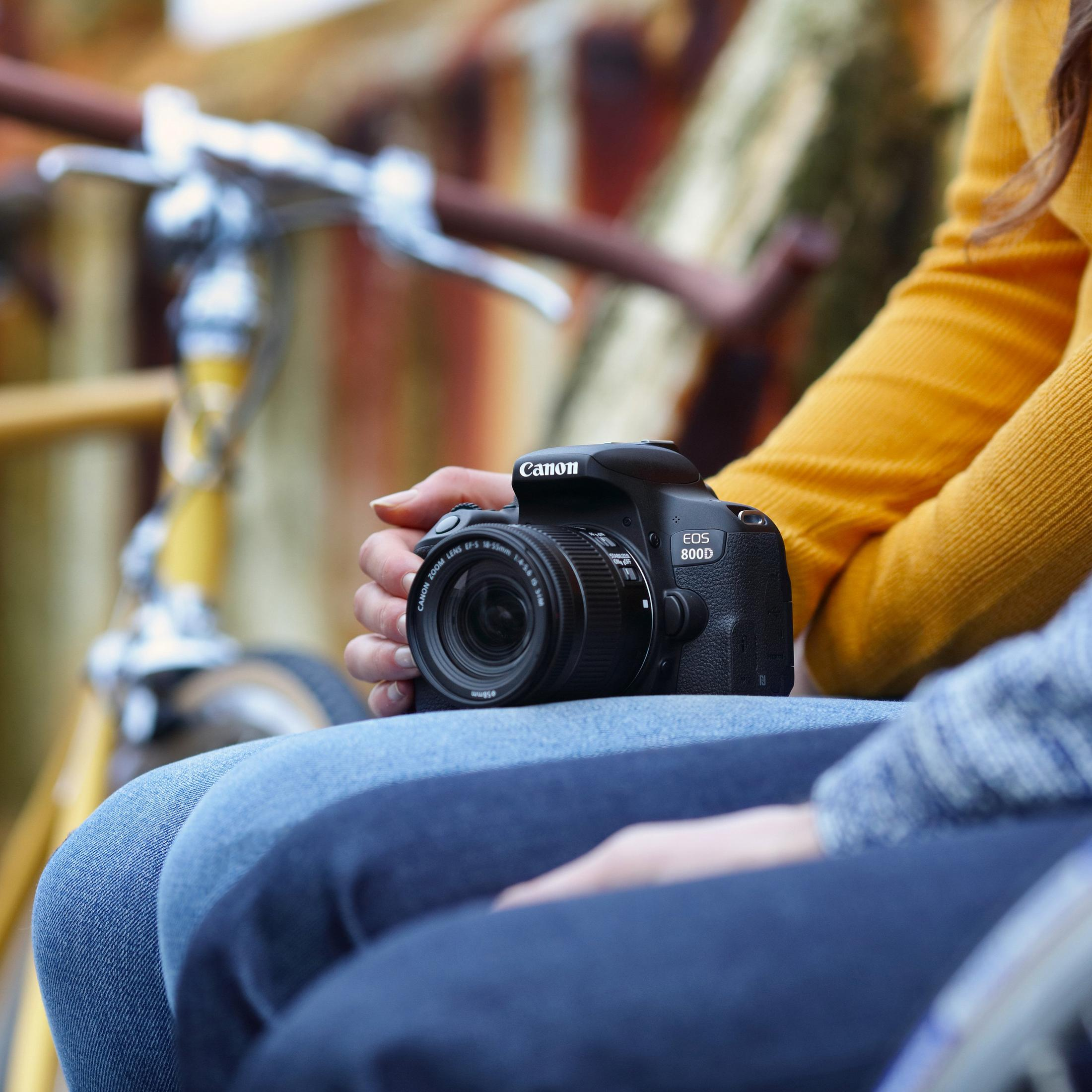 Una persona tiene una fotocamera Canon EOS 800D nera. Una bicicletta sullo sfondo.