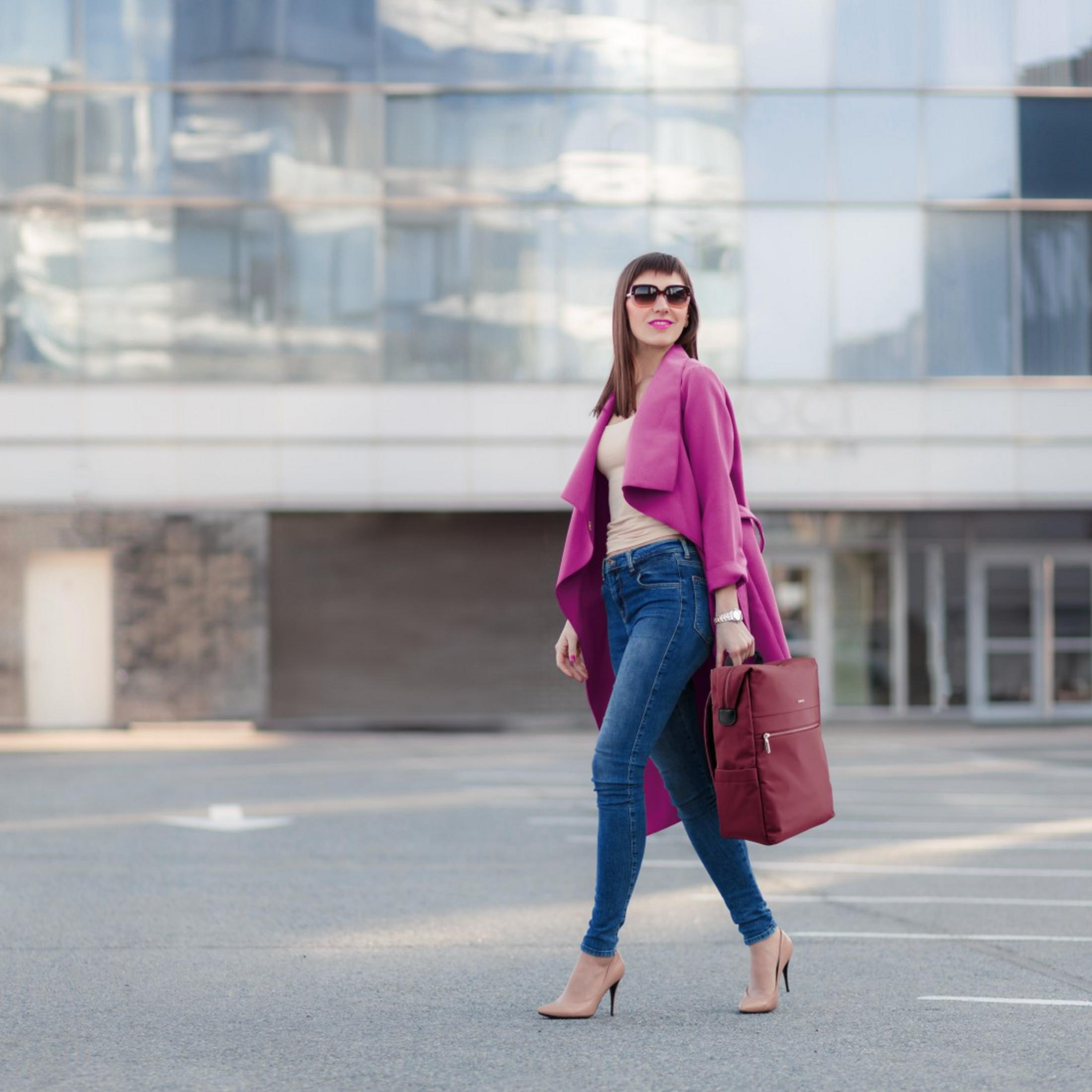 Una donna in jeans e cappotto rosa cammina davanti a un edificio moderno.