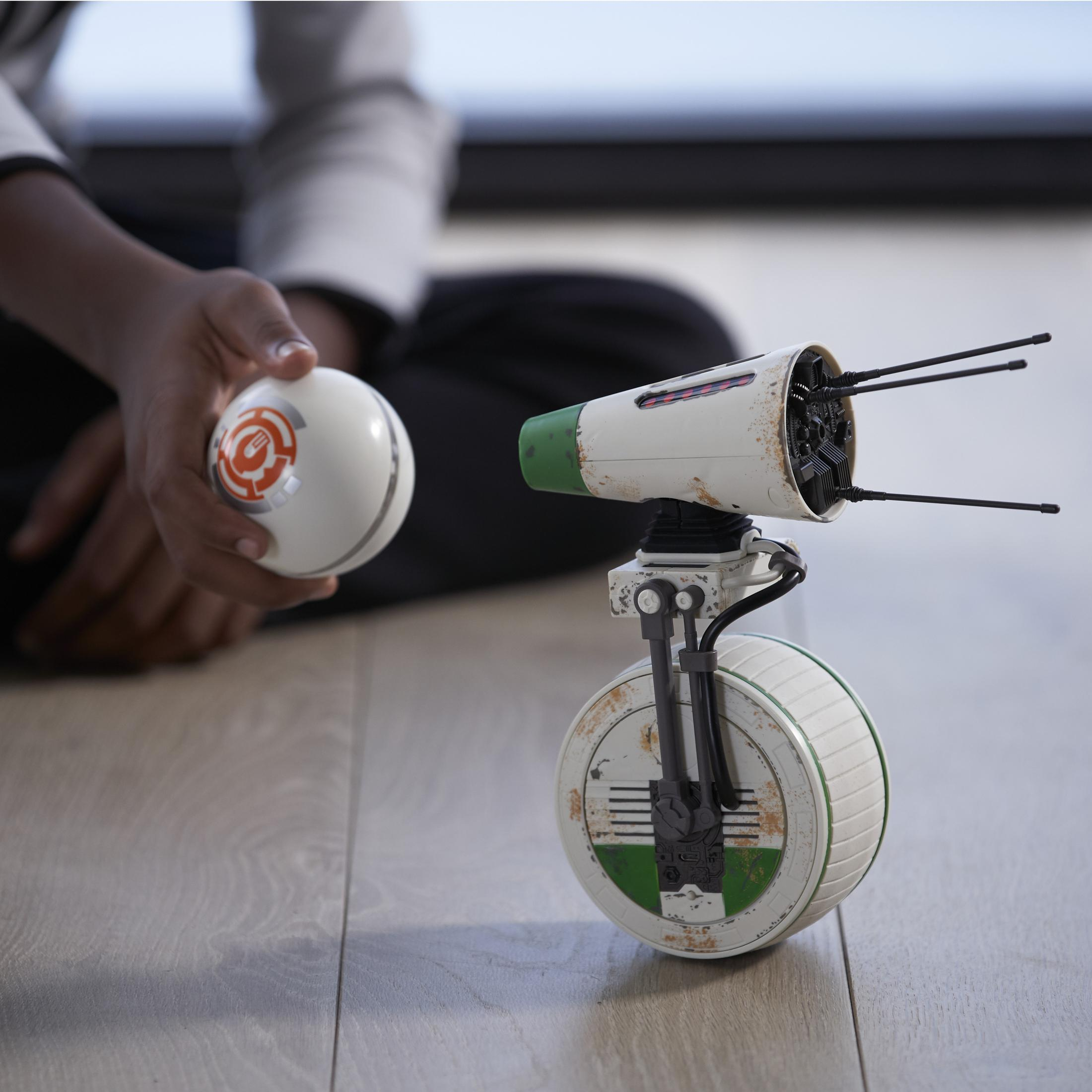 Kinderhand hält weißen Ball mit orangen Markierungen neben weiß-grünem Roboter auf Holzboden.