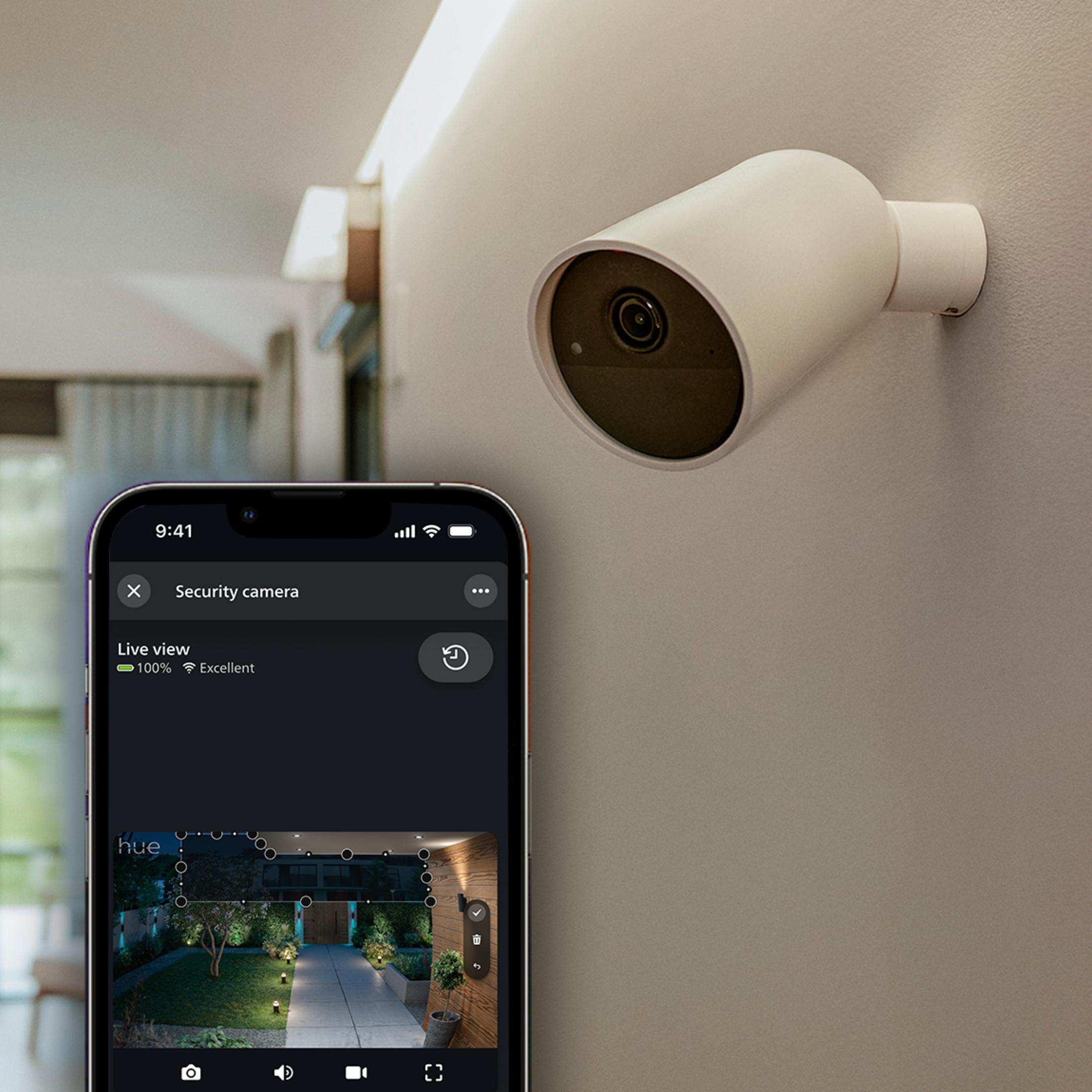 Una cámara de seguridad blanca montada en una pared junto a un smartphone mostrando vista en vivo.